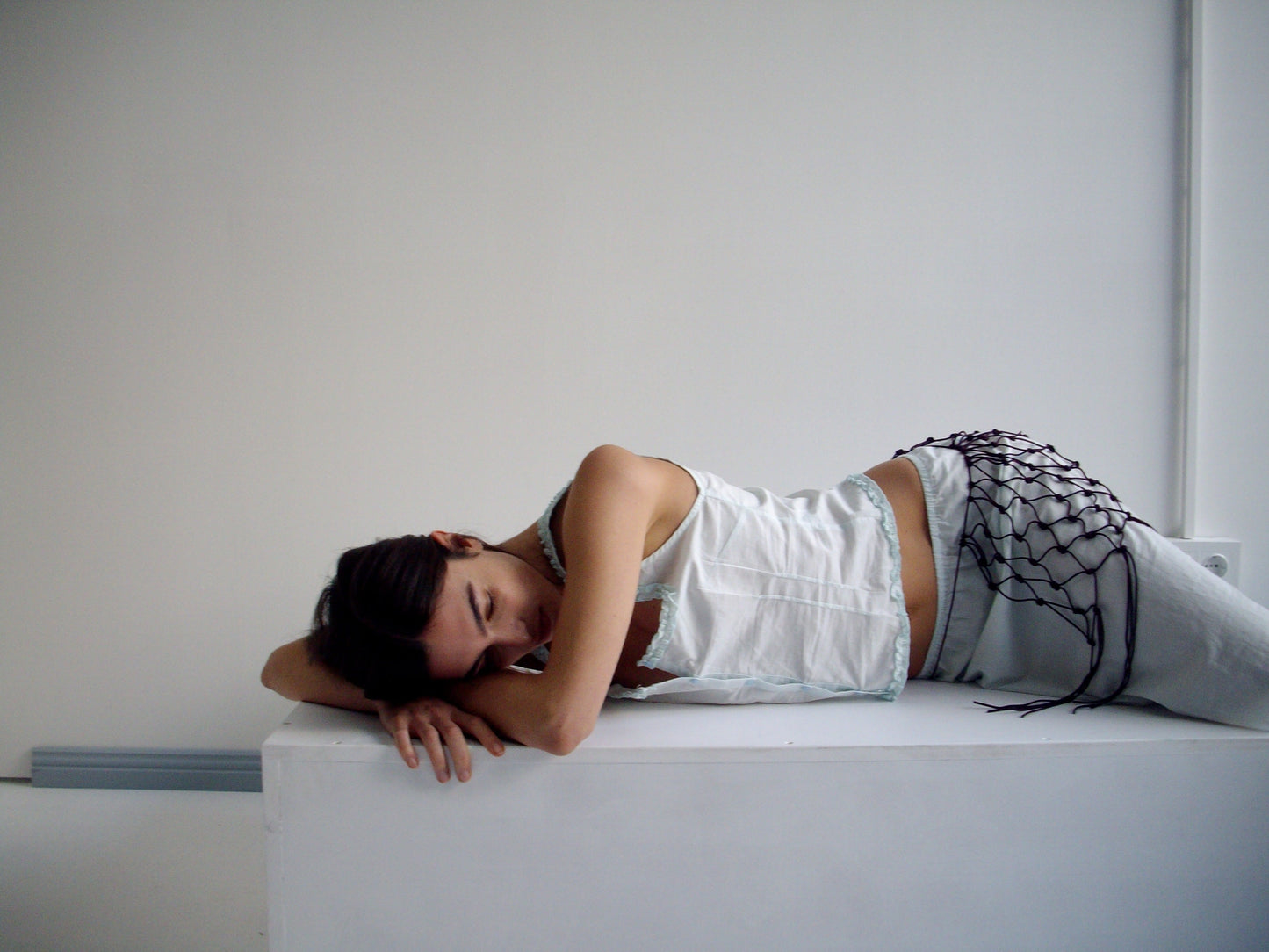 A woman in light clothing and a burgundy Macrame Belt lies on her side on a white surface in a minimalist, light-filled room, resting her head on her arm and looking downward.