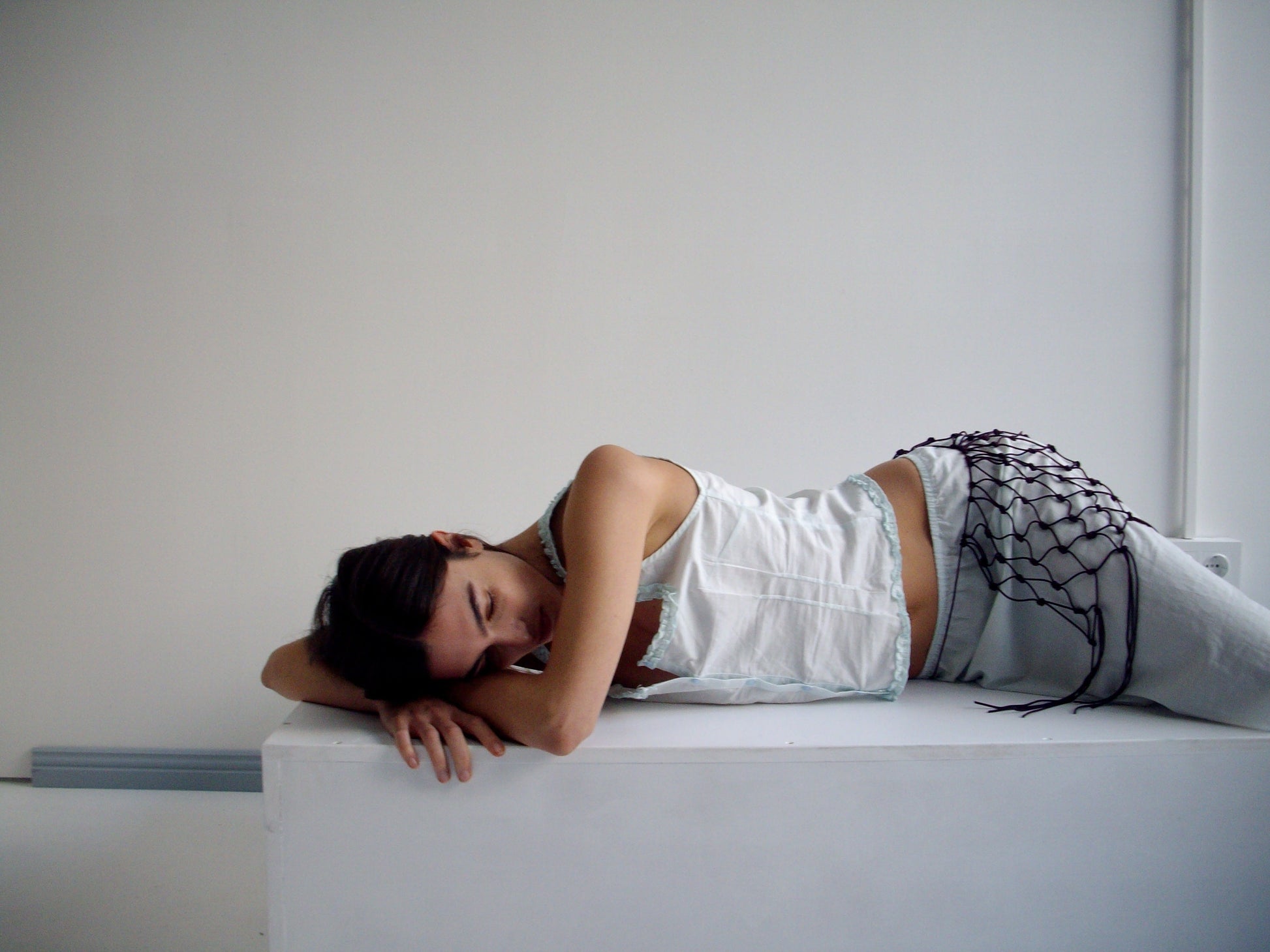 A woman in light clothing and a burgundy Macrame Belt lies on her side on a white surface in a minimalist, light-filled room, resting her head on her arm and looking downward.