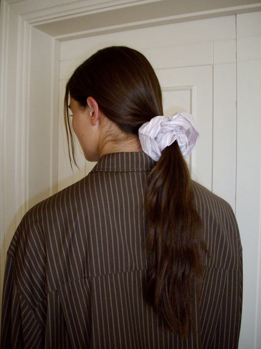 A person with long brown hair tied in a low ponytail by a Scrunchie stands facing away from the camera, wearing a brown pinstriped shirt in front of a white door.