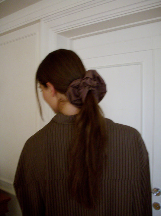 A person with long brown hair tied in a ponytail with a large, dark Scrunchie is wearing a brown pinstriped shirt and standing with their back to the camera in front of a white door.