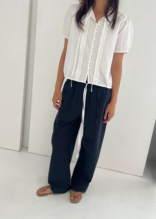 A person stands indoors against a plain white background wearing the Double Frill Top, a short-sleeved white blouse with ribbon ties and ruffle details, paired with loose black pinstriped pants and tan sandals. The person’s face is not visible.