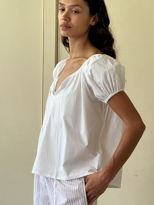 A woman stands indoors against a neutral background, wearing the One Panel Top—a loose white short-sleeve blouse with puffed sleeves—and light-colored striped pants, gazing slightly to the side with one hand in her pocket.