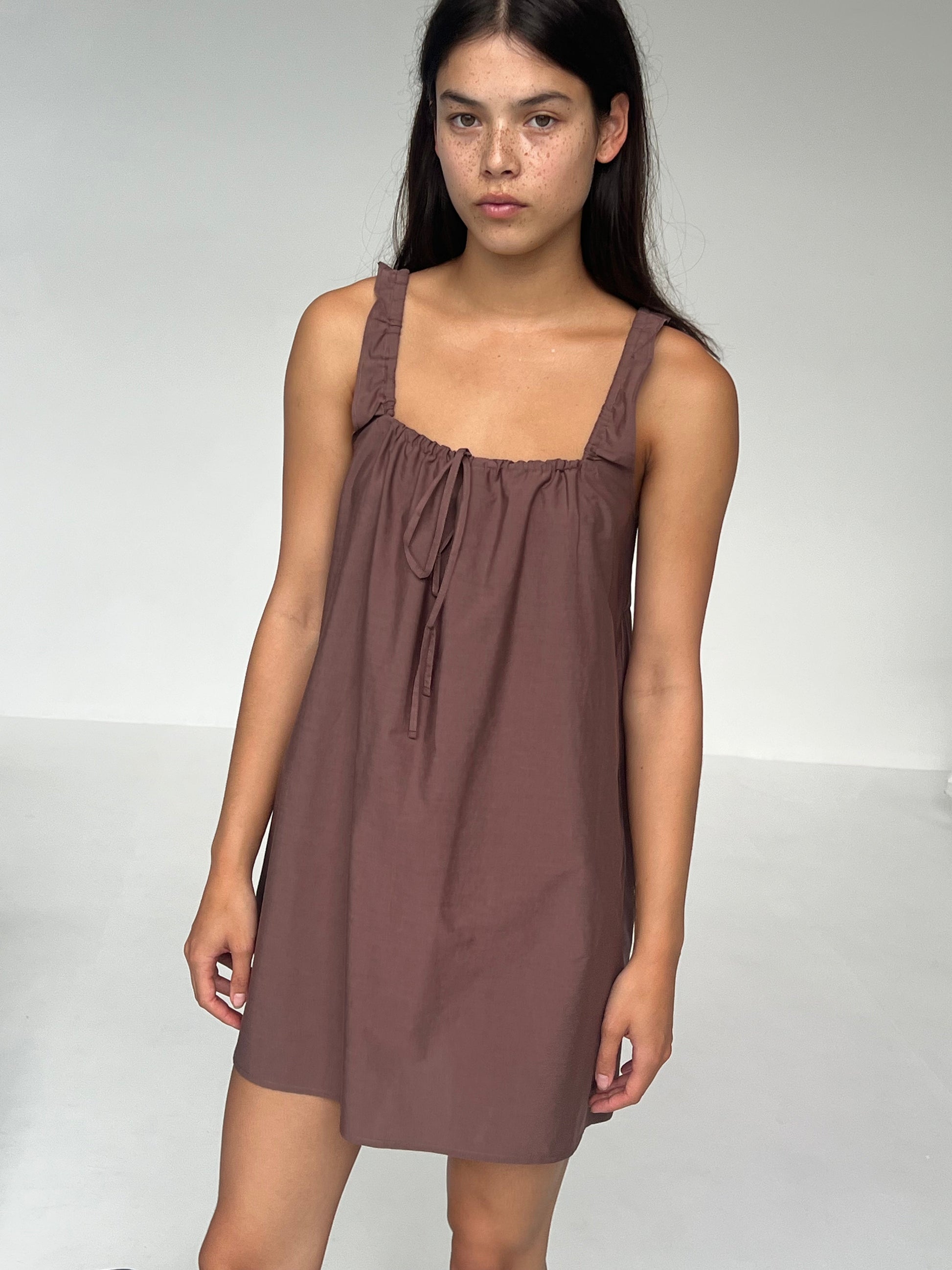 A young woman with long dark hair stands against a white background wearing The Bow Dress - Cedar, a sleeveless brown mini dress featuring ruffled straps and a tie neckline. She has a neutral expression.