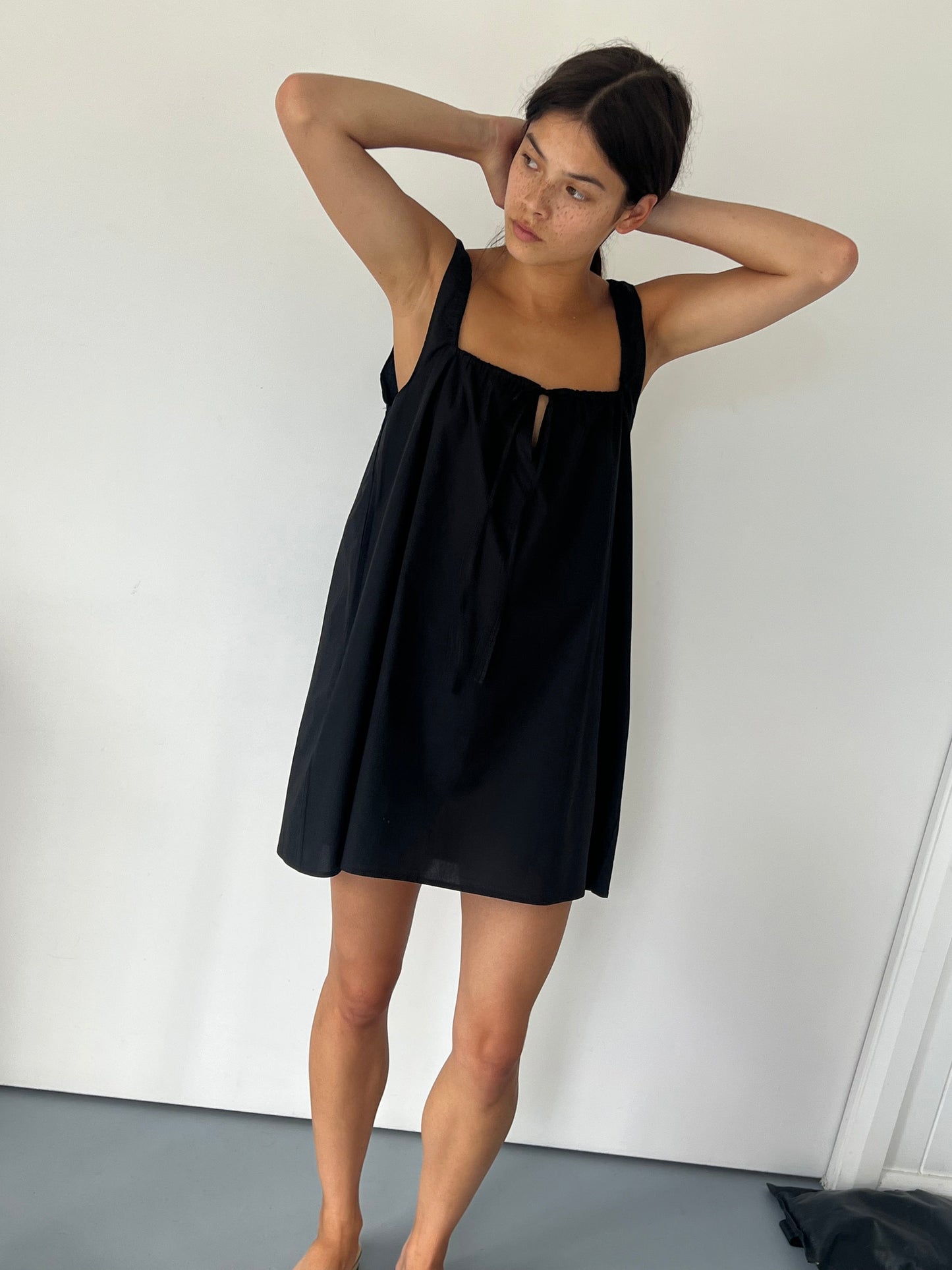 A woman stands barefoot indoors against a white wall, wearing The Bow Dress—a loose, sleeveless black piece. Her dark hair is pulled back as she raises her arms to touch the back of her head, gazing off to the side.