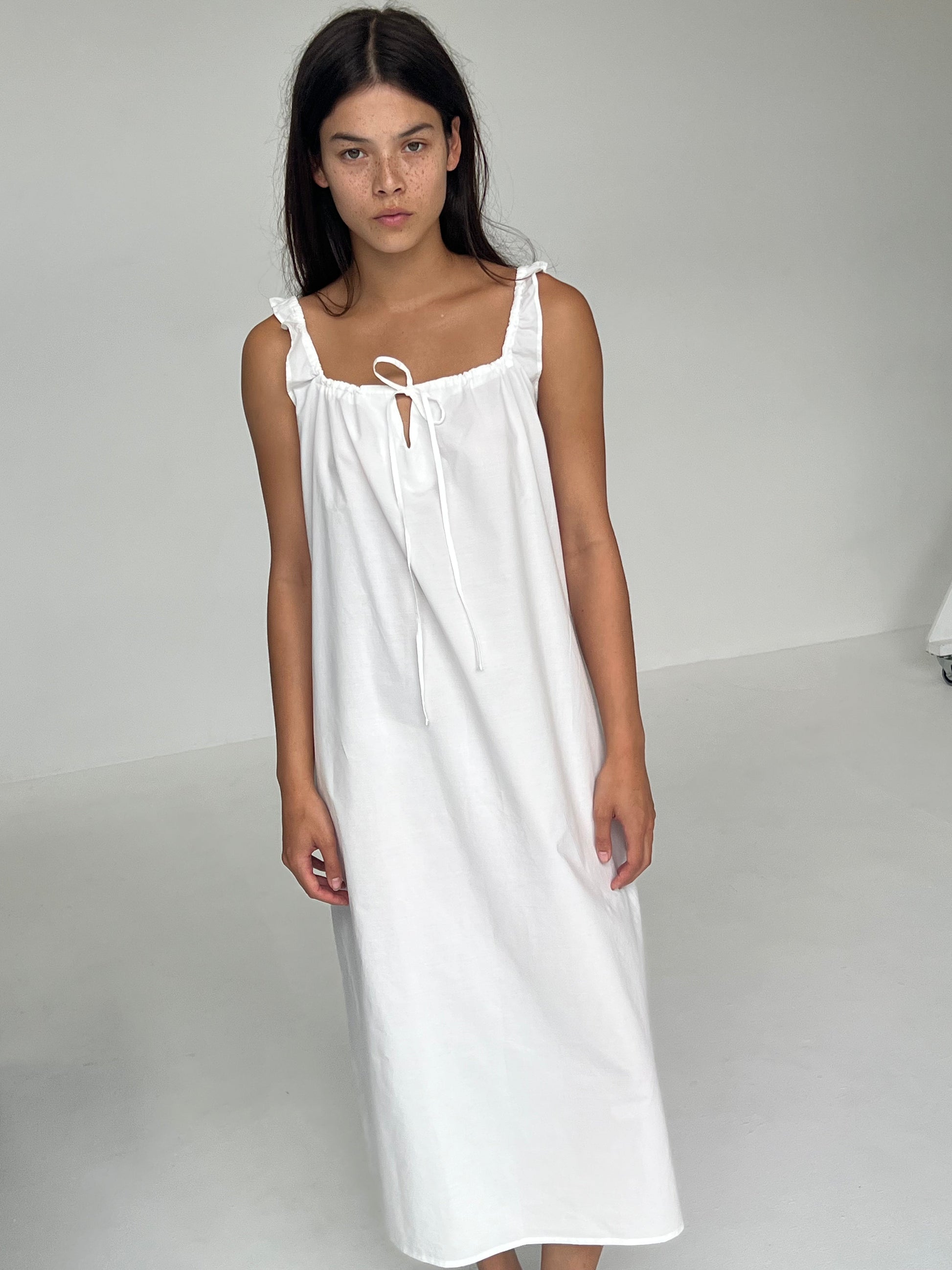 A person with long dark hair stands indoors against a plain light background, wearing The Paper Dress - White from Deiji Studios: a sleeveless, ankle-length white maxi dress with ruffle details and a tie at the neckline.