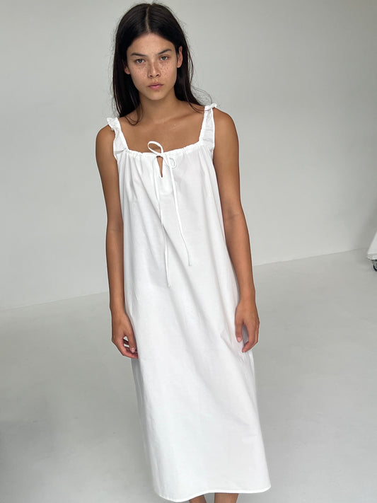 A person with long dark hair stands indoors against a plain light background, wearing The Paper Dress - White from Deiji Studios: a sleeveless, ankle-length white maxi dress with ruffle details and a tie at the neckline.