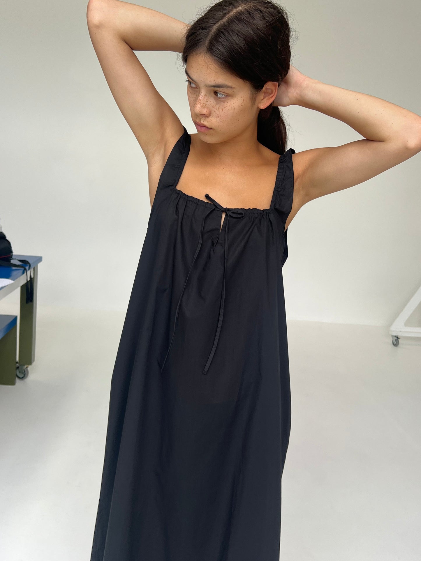 A person with long dark hair stands indoors, wearing The Paper Dress—a loose black sleeveless piece. They look to the side while adjusting their hair, set against a plain and minimal background.