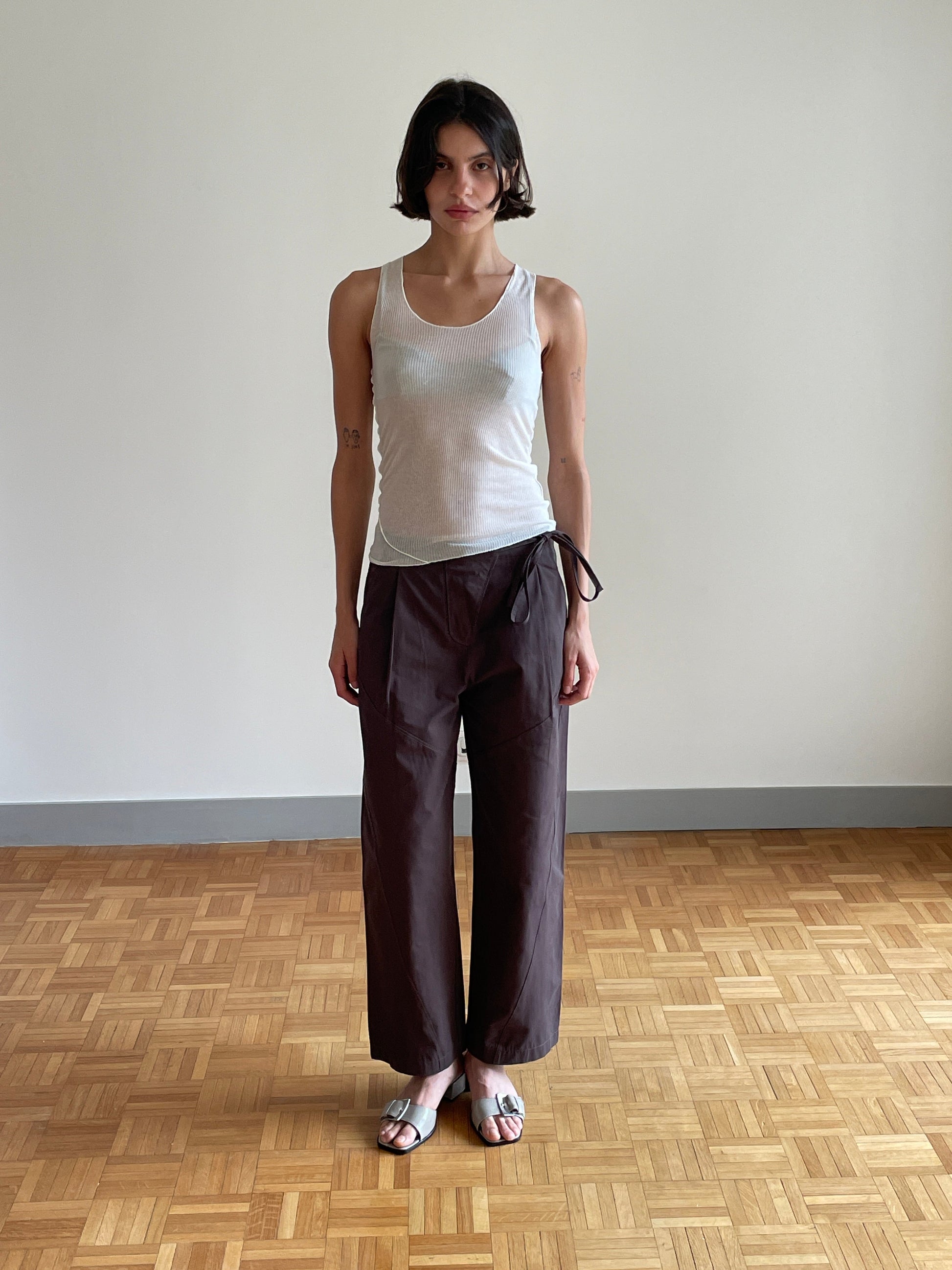 A person stands indoors on a wooden floor, wearing a sheer white tank top, Flag Tie Pant in dark color with side tie, and white sandals, facing forward against a plain light-colored wall.