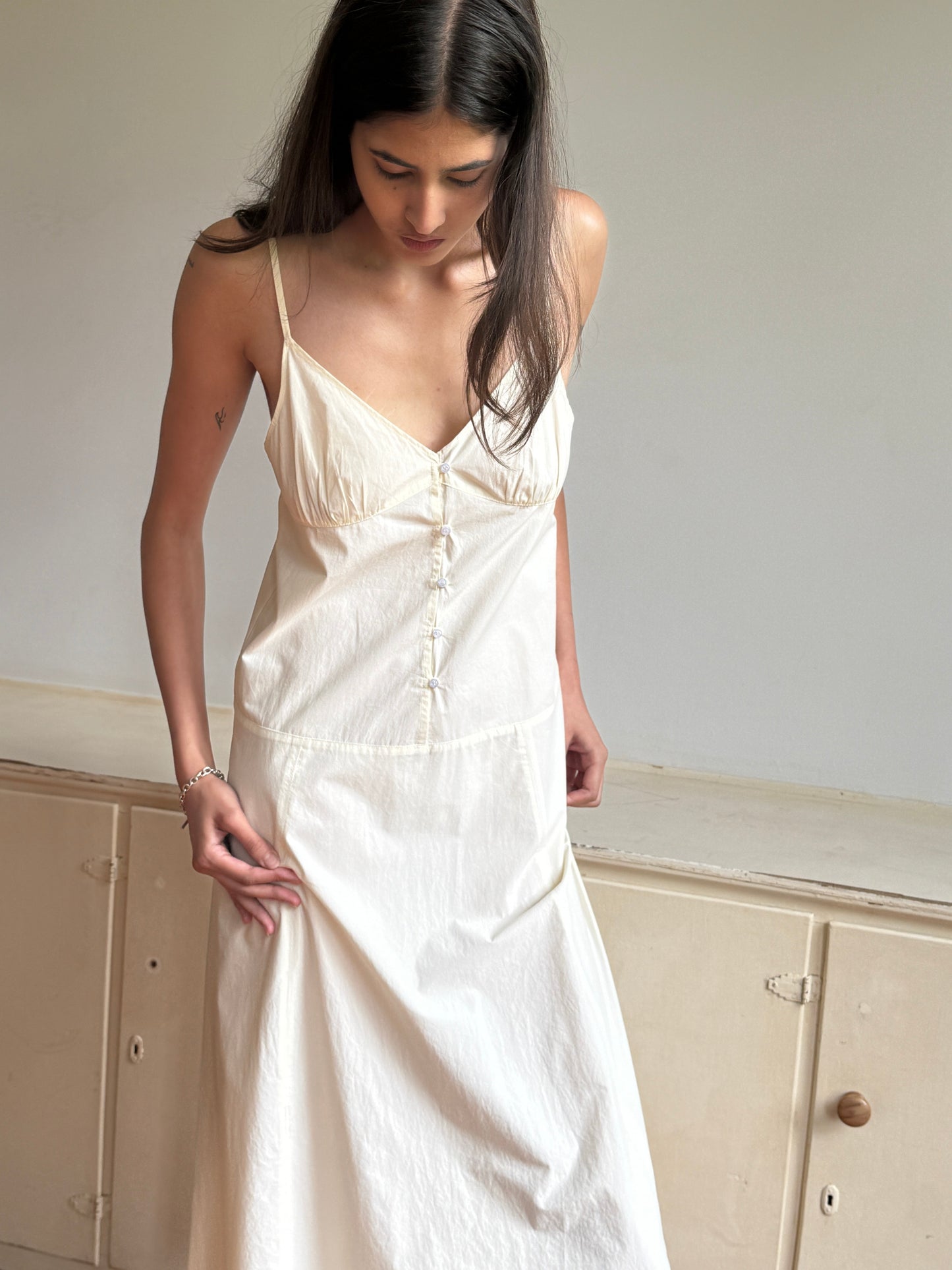 A woman with long dark hair wears the 0 Double Seam Dress - Butter White, looking down as she gently holds the fabric in a softly lit, minimalist room with light-colored furniture.