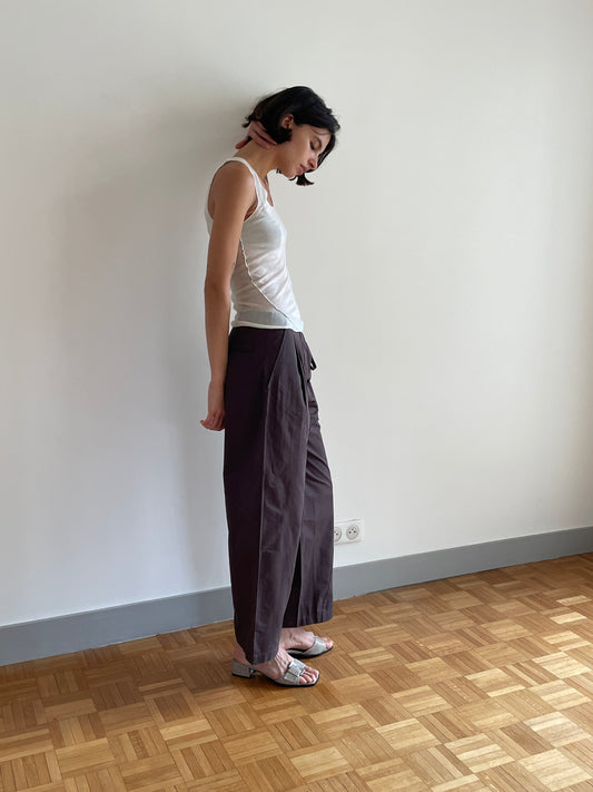 A woman with short dark hair stands sideways against a white wall, wearing a Deiji Studios white sleeveless top, Flag Tie Pant in dark brown, sandals, and a pink scarf. She stands on a wooden parquet floor.