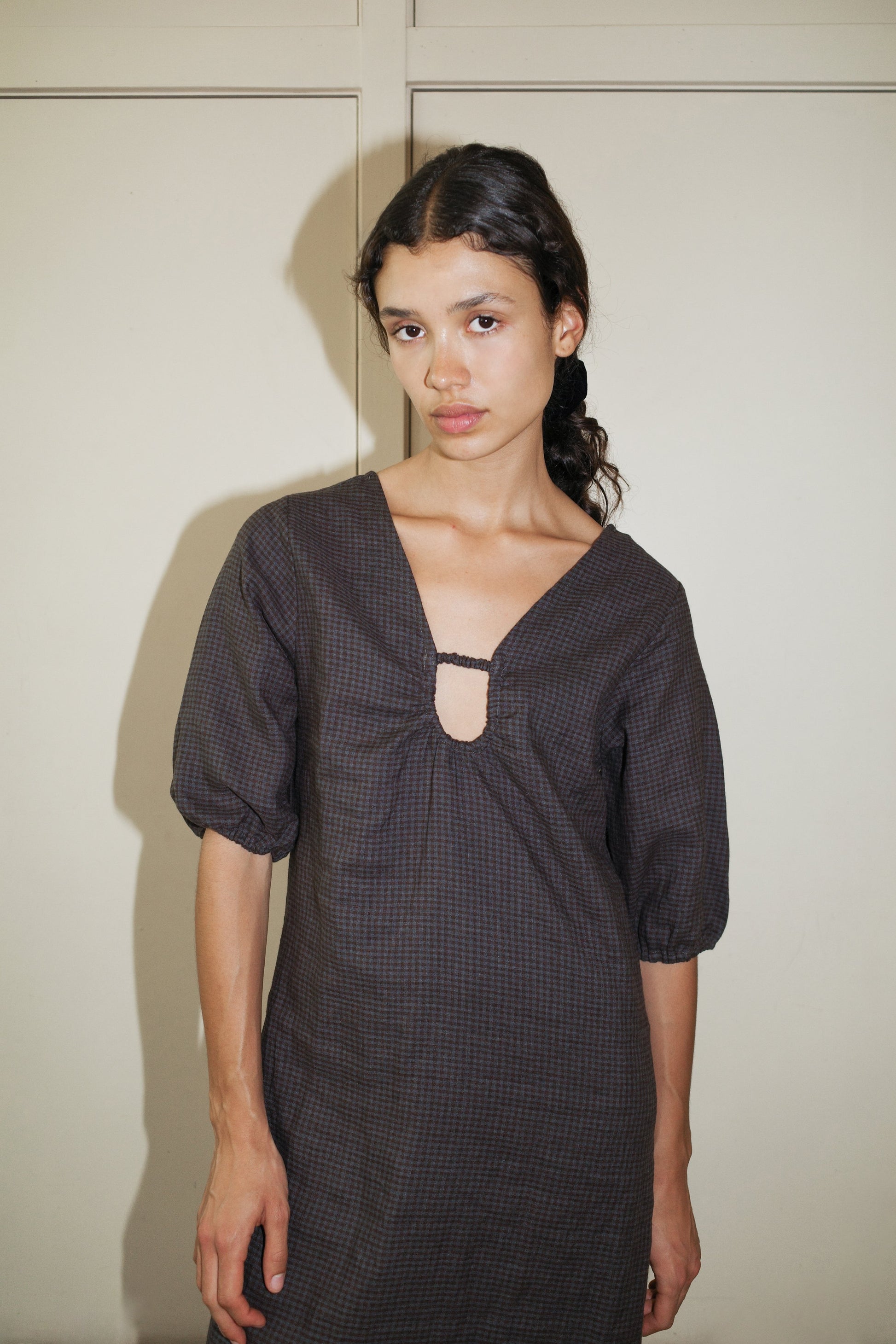 A young woman with dark hair in a low braid wears the Squared Dress - Slate Check by Deiji Studios, featuring puffed sleeves and a keyhole neckline in organic linen, standing against a plain light wall and looking at the camera.