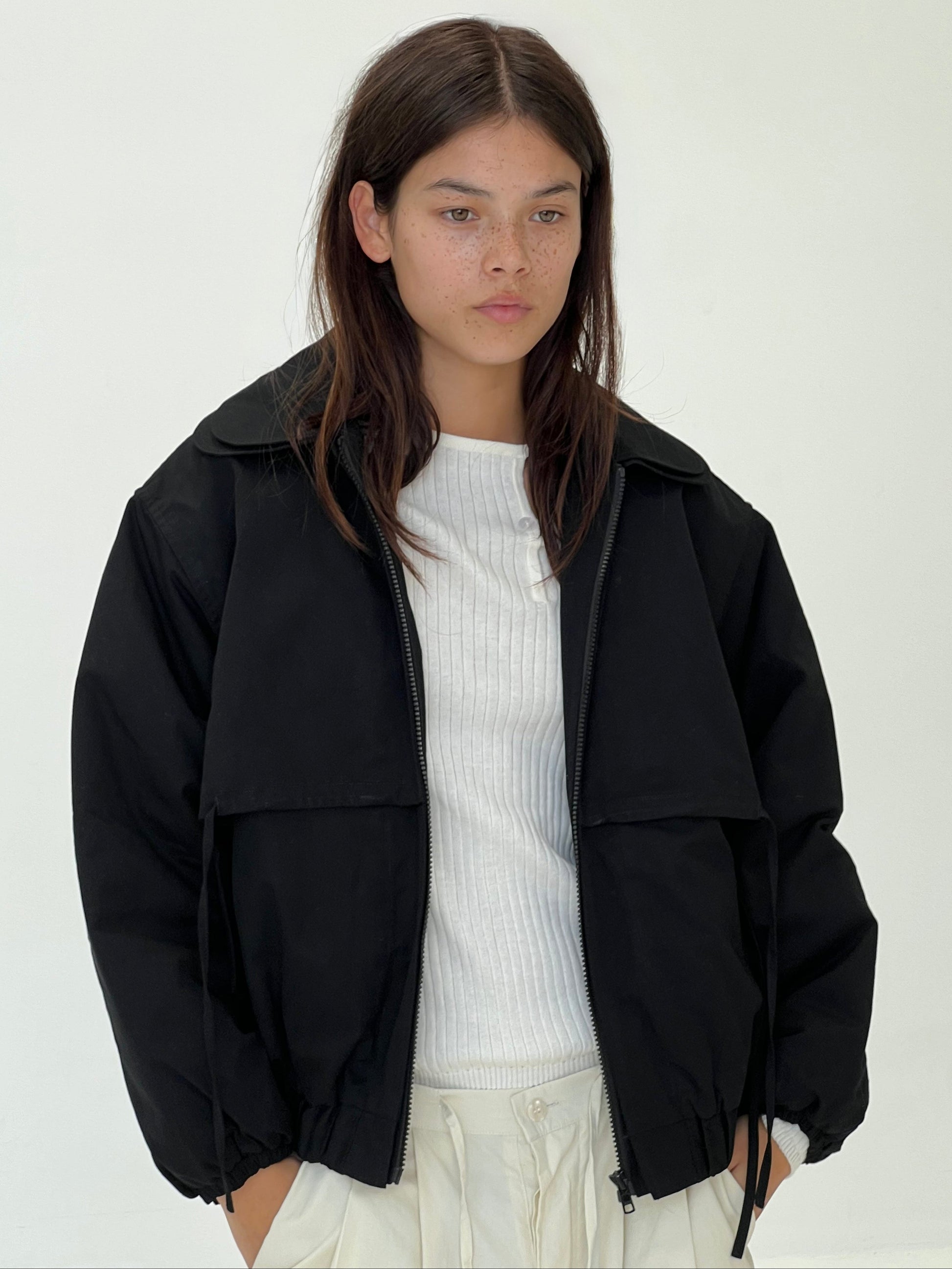 A young woman with straight brown hair and light skin stands against a plain background, wearing the Double Layered Jacket - Black over a white ribbed shirt and white pants. Hands in her pockets, she gazes ahead calmly.