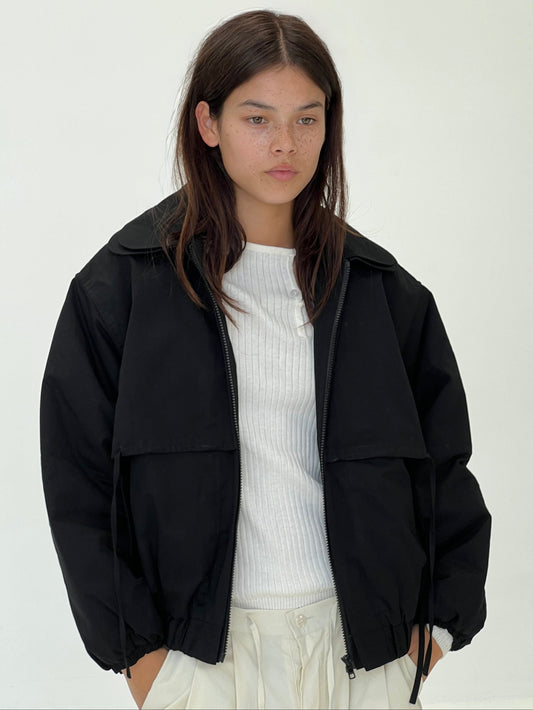 A young woman with straight brown hair and light skin stands against a plain background, wearing the Double Layered Jacket - Black over a white ribbed shirt and white pants. Hands in her pockets, she gazes ahead calmly.