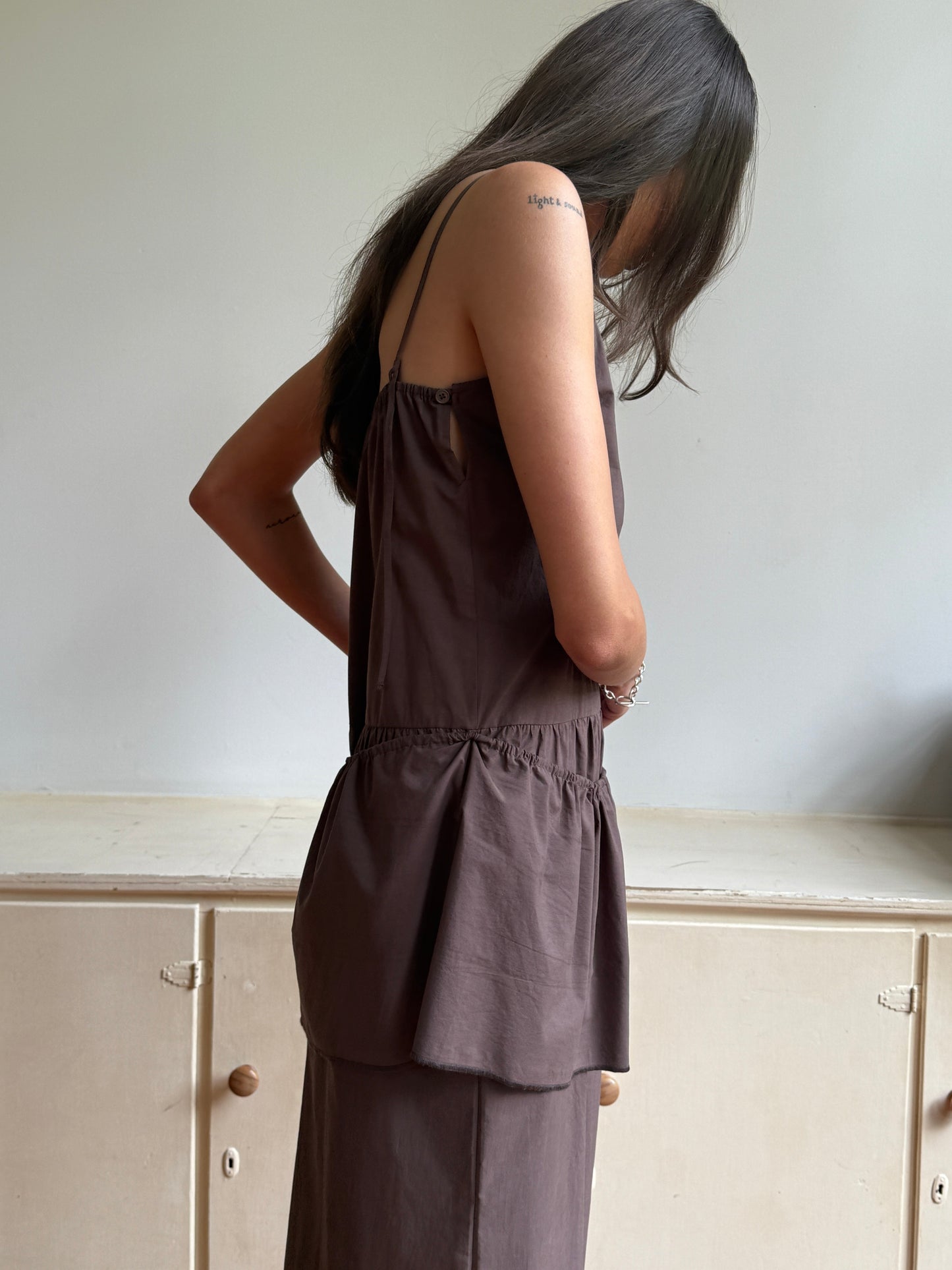 A woman with long dark hair stands indoors by a light cabinet, looking down with arms crossed, wearing the 0 Peplum Dress in Chocolate—a sleeveless organic cotton piece.