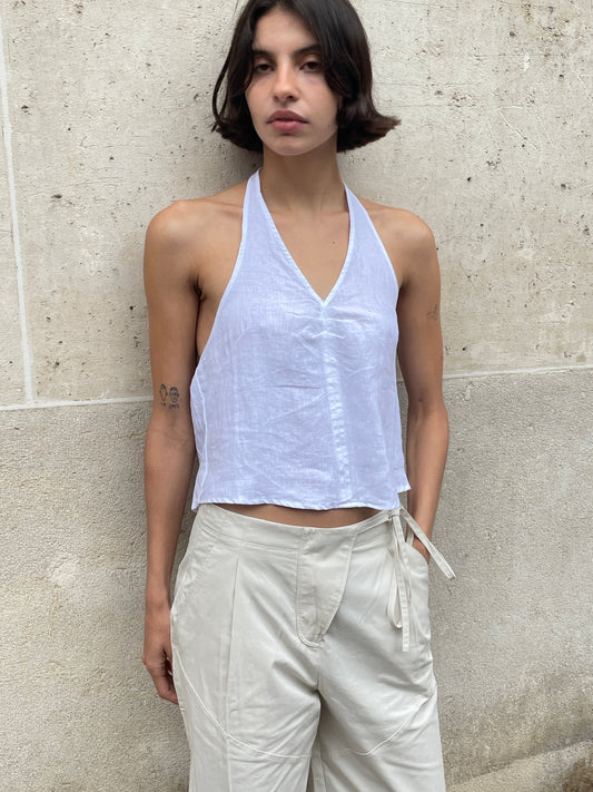 A person with short dark hair stands against a beige wall, wearing a sleeveless white halter top and the Flag Tie Pant - Off White, featuring a relaxed straight leg and side flag tie detail, looking slightly to the side with a neutral expression.