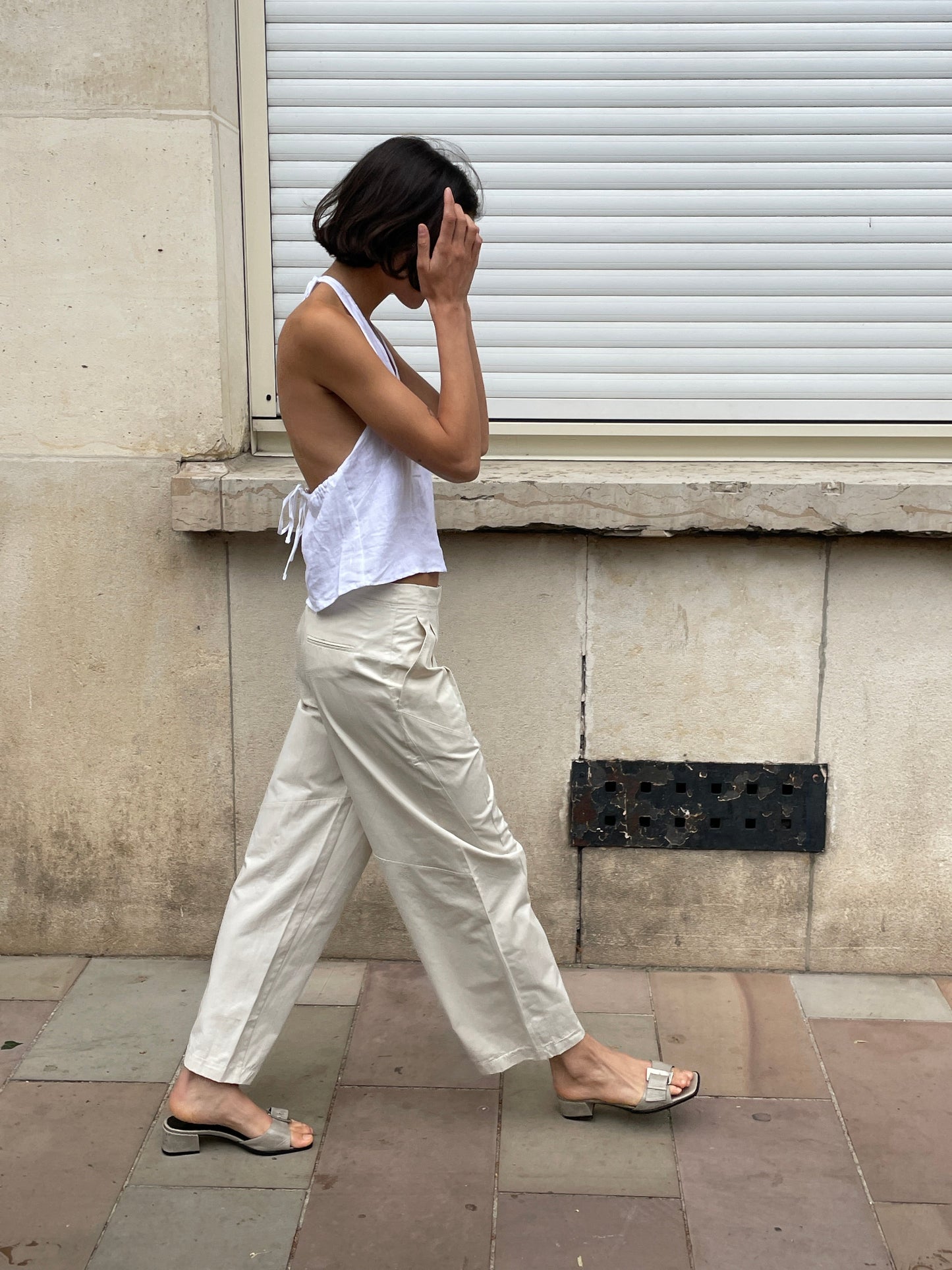A person with short dark hair, wearing a sleeveless white top and the Flag Tie Pant - Off White, walks along a sidewalk in sandals, covering their face with one hand beside a beige building with a closed shutter.