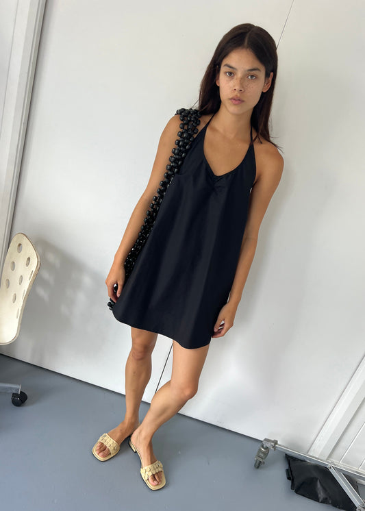 A woman with long dark hair wears the Round Halter Dress - Black from Deiji Studios and beige slide sandals, standing indoors near a white wall. She carries a black beaded bag, with a white chair beside her.