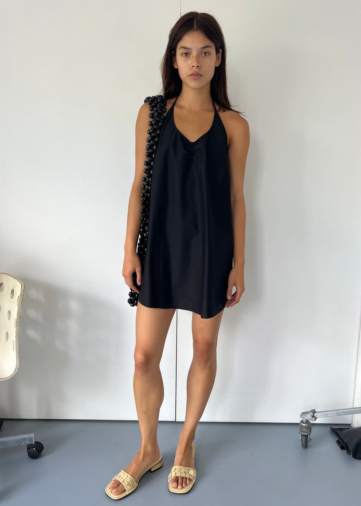 A woman with long dark hair stands indoors against a white wall, wearing the Round Halter Dress - Black. She pairs it with beige woven sandals and a black beaded shoulder bag. A white chair and metal clothing rack are nearby.