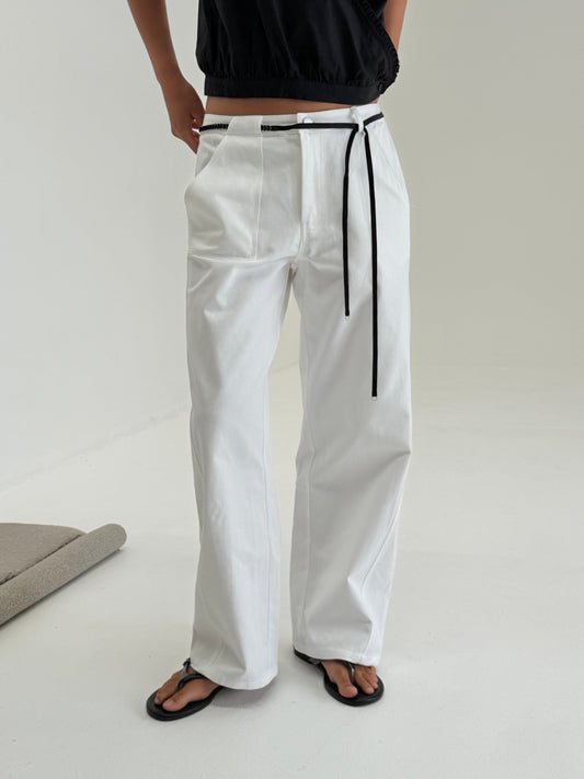 A person stands indoors wearing Pocket Jean, a black short-sleeve top, and black flip-flops. Their hands are in their pockets, and their head is not visible.