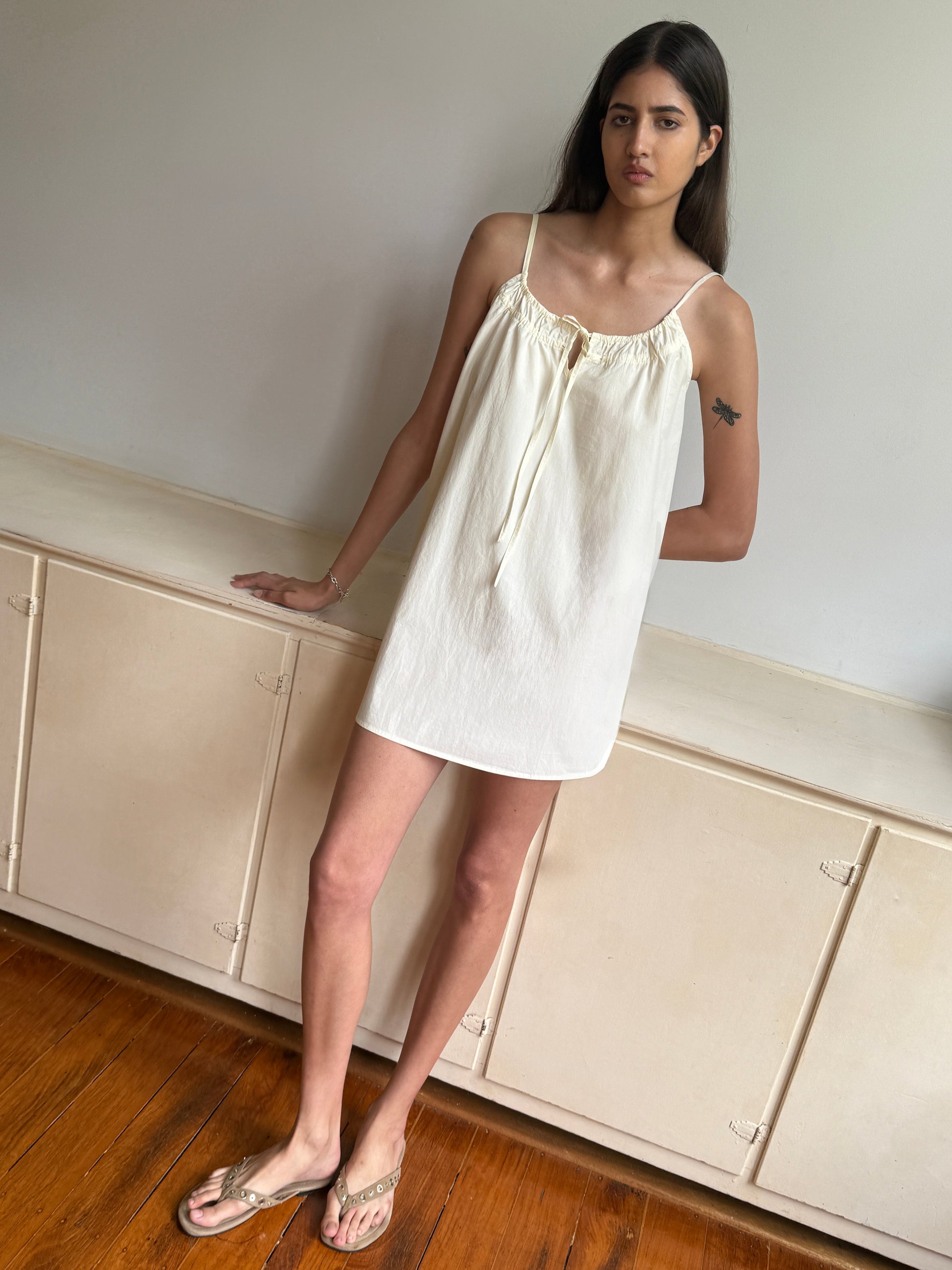 A woman with long dark hair and a tattoo on her upper left arm wears the Light Dress - Butter White by 0, paired with clear sandals, standing indoors against a cream cabinet on a wooden floor. She has a neutral expression.