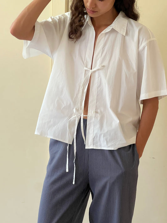 A person wears The Tie Up Shirt, a loose white short-sleeve top tied at the chest, styled with blue pinstripe pants and standing with one hand in their pocket against a plain background.