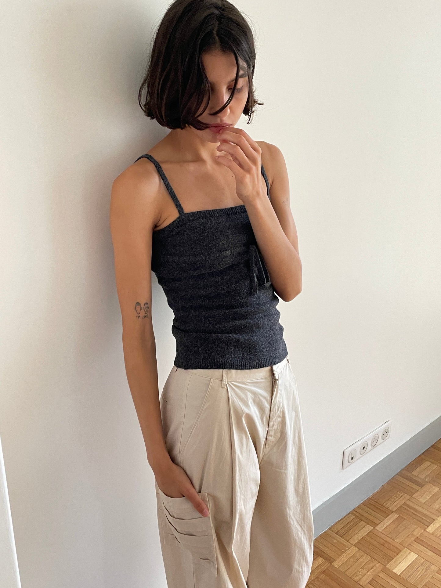 A person with short dark hair stands indoors against a white wall, wearing a dark sleeveless top and the Gathered Pocket Pant - Off White, looking down with one hand near their face and the other in their pocket.