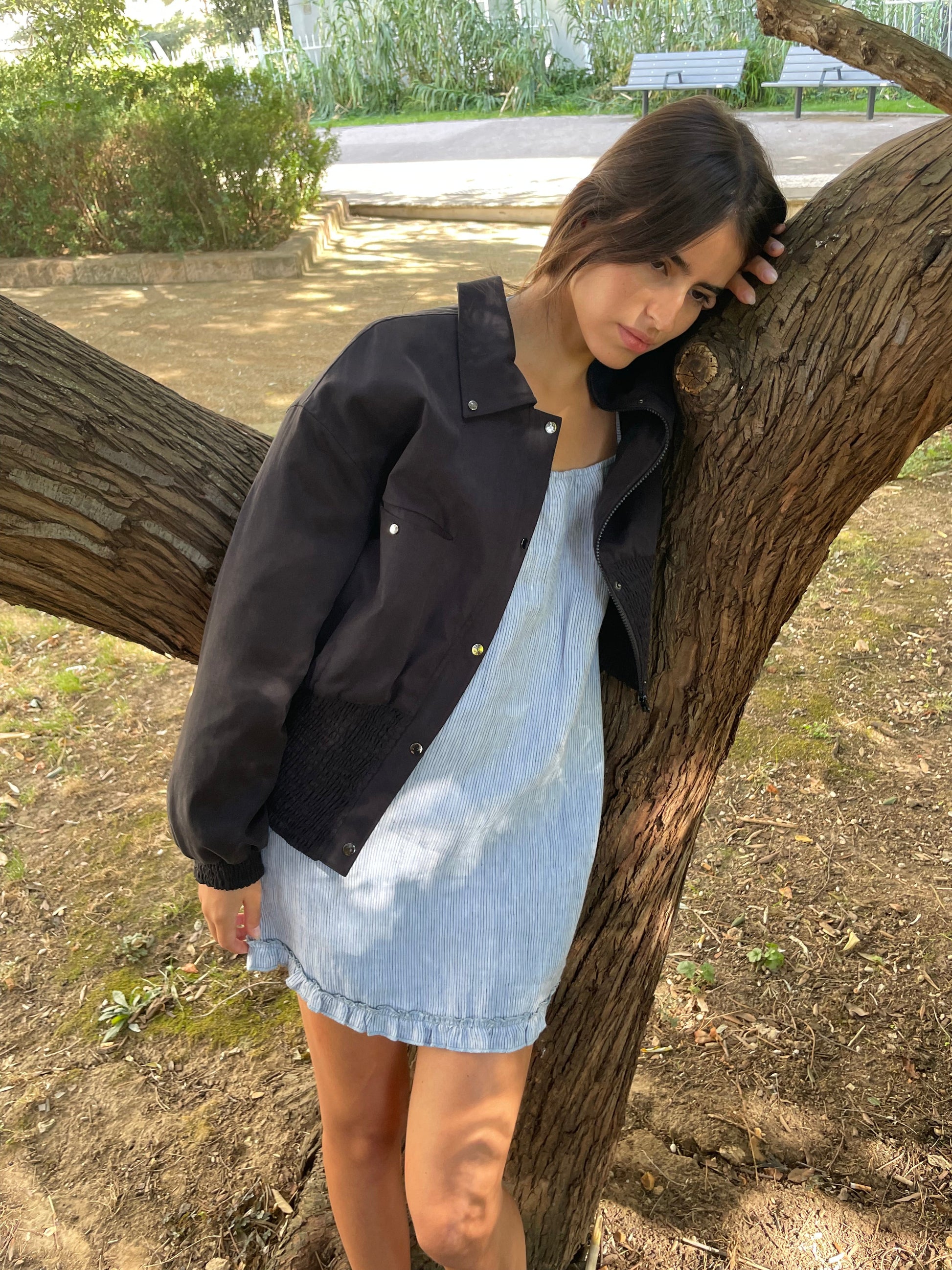 A woman in a light blue dress and the Shirring Jacket - Slate leans thoughtfully against a tree branch in a sunlit park, looking down with greenery and a bench in the background.