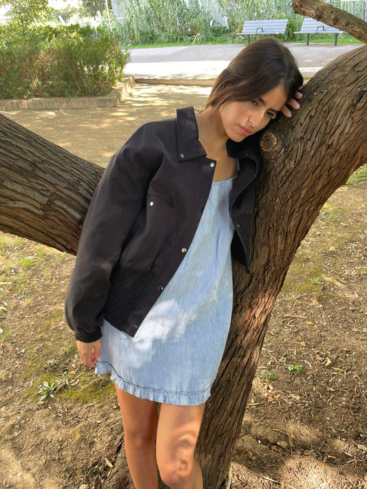 A woman in a light blue dress and the Shirring Jacket - Slate leans thoughtfully against a tree branch in a sunlit park, looking down with greenery and a bench in the background.
