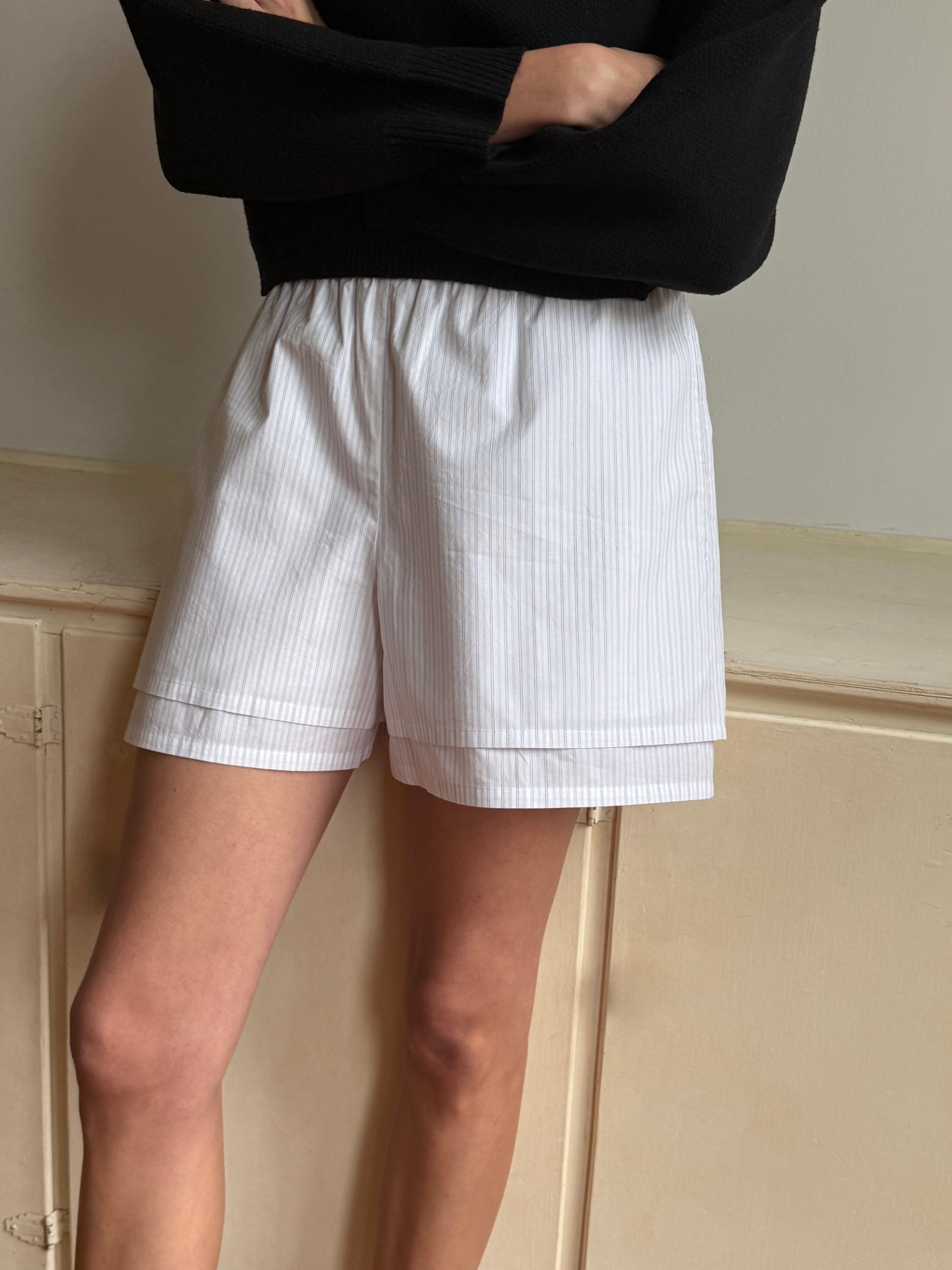 Someone stands with arms crossed in front of a cream wall, wearing a black long-sleeve top and the Layered Short - Pacific Stripe, featuring white fabric with subtle stripes and an elastic waist. Their head is not visible in the image.