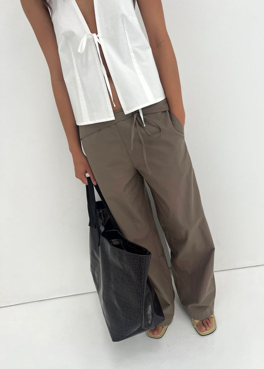 Wearing a white sleeveless top with a front tie and the Belted Pant - Clay from Deiji Studios, a person stands against a white background holding a large black tote bag. Only their torso and legs are visible.
