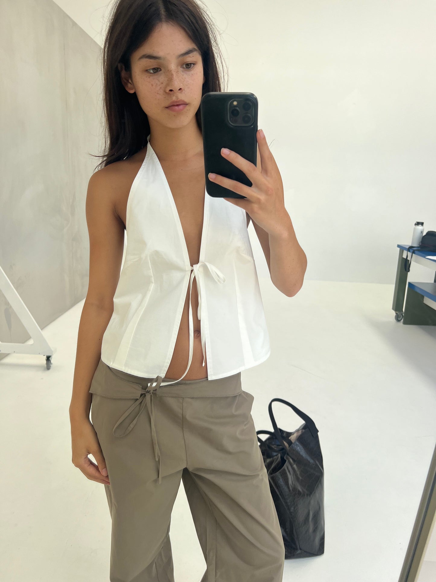 A person with long dark hair and freckles takes a mirror selfie in a minimal studio, wearing a white halter top and the Belted Pant - Clay. A large black bag is on the floor beside them.