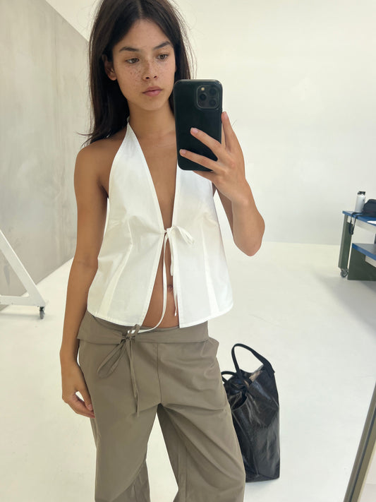 A person with long dark hair and freckles takes a mirror selfie in a minimal studio, wearing a white halter top and the Belted Pant - Clay. A large black bag is on the floor beside them.