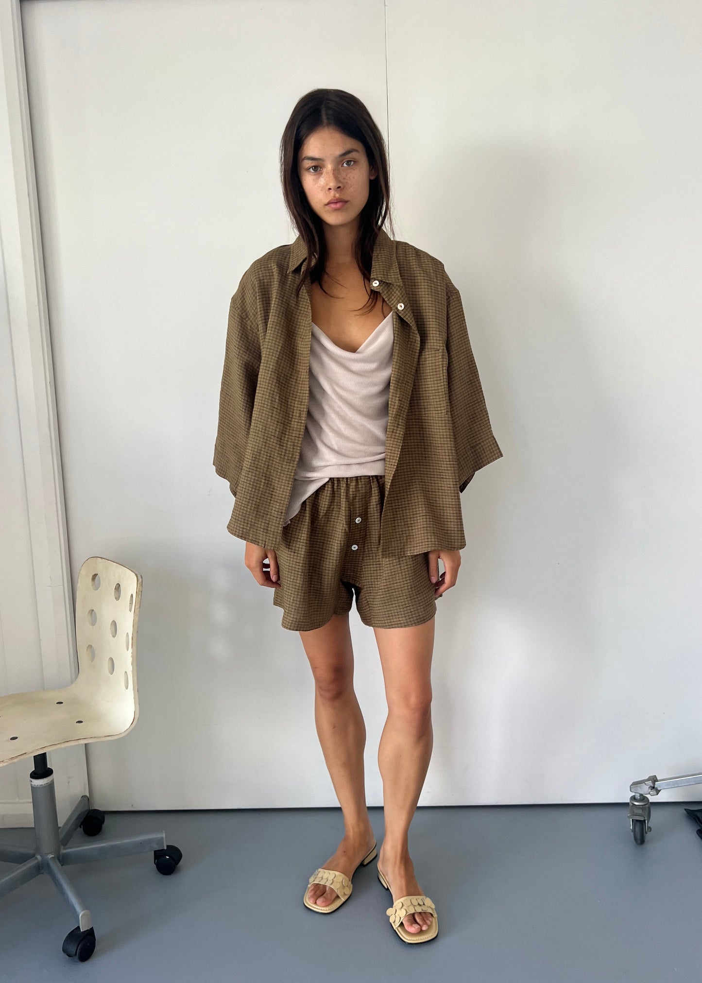 A model wears the 03 Set - Pine Check by Deiji Studios, featuring a French linen shirt and matching shorts, paired with a beige camisole and neutral woven slide sandals, standing against a white wall.