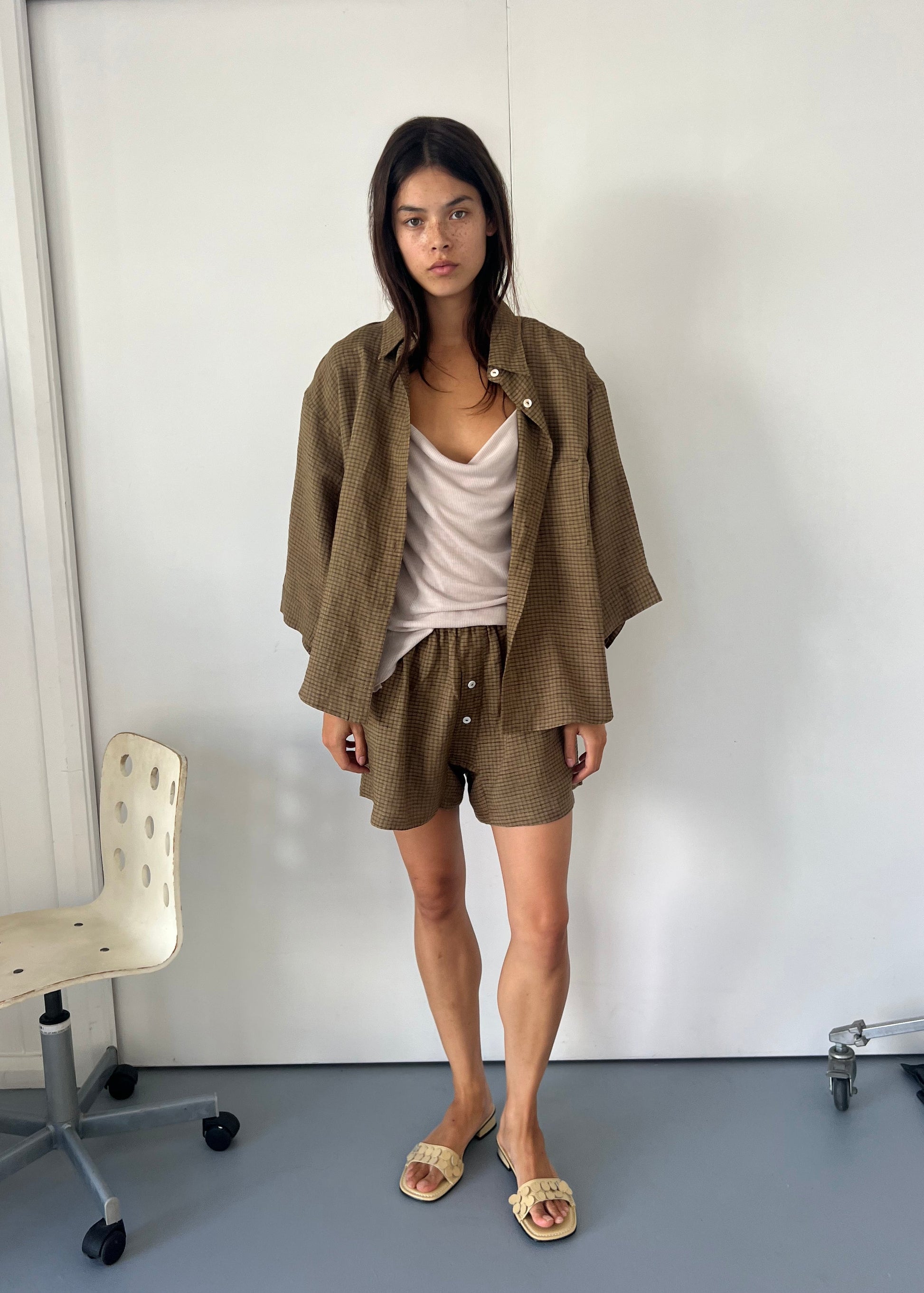 A model wears the 03 Set - Pine Check by Deiji Studios, featuring a French linen shirt and matching shorts, paired with a beige camisole and neutral woven slide sandals, standing against a white wall.