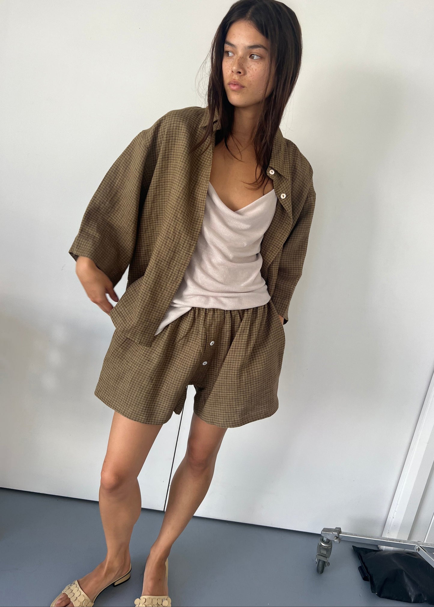 A woman with long dark hair and a neutral expression wears the 03 set—loose brown checked shorts and shirt—over a light beige top. Barefoot, she stands on a light floor against a plain white wall.