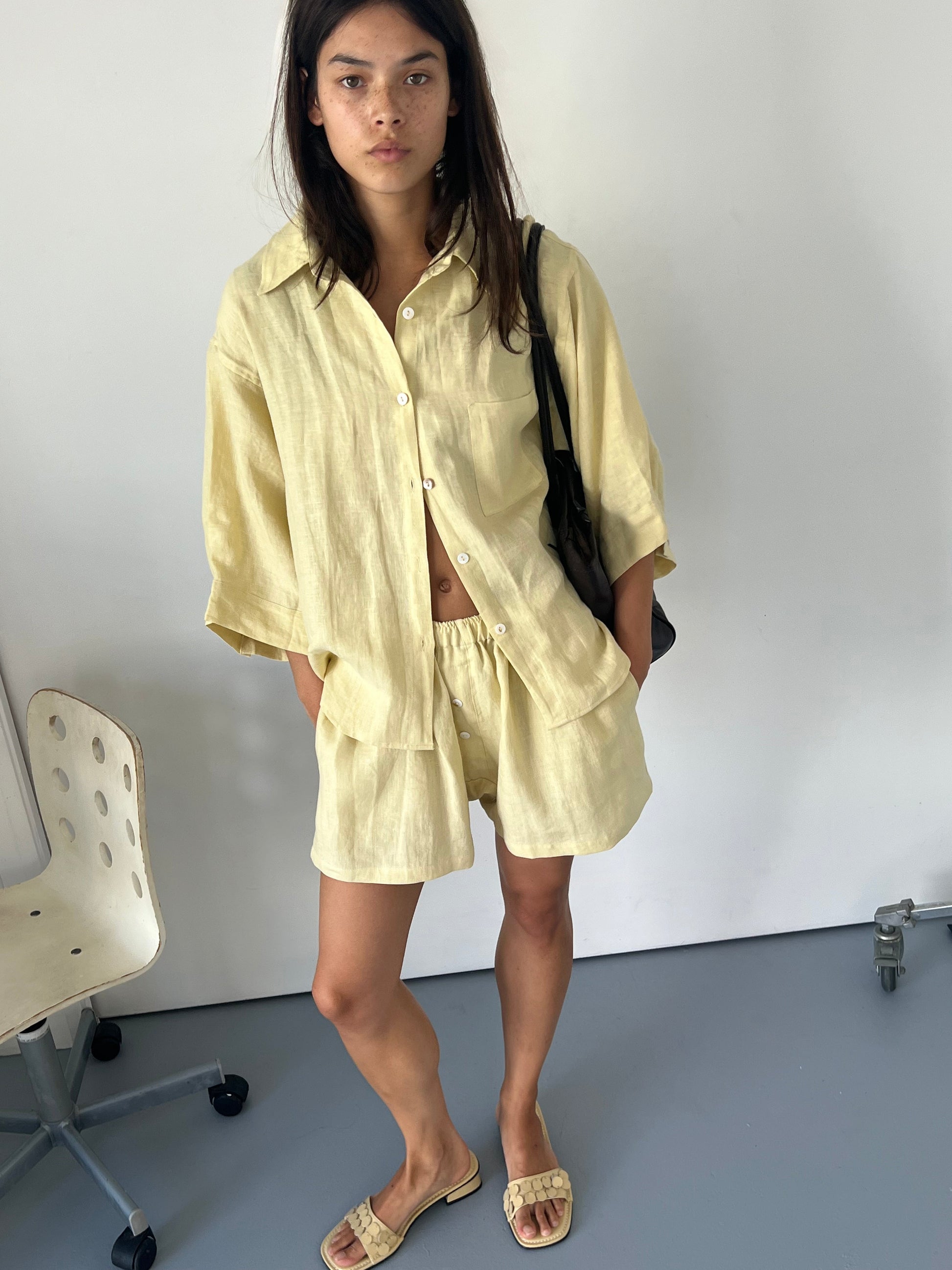 A woman with long dark hair wears the 03 Set - Butter Yellow, featuring a light yellow organic linen shirt and matching boxer shorts. She carries a black bag, wears beige slide sandals, and stands indoors beside a white chair on a gray floor.
