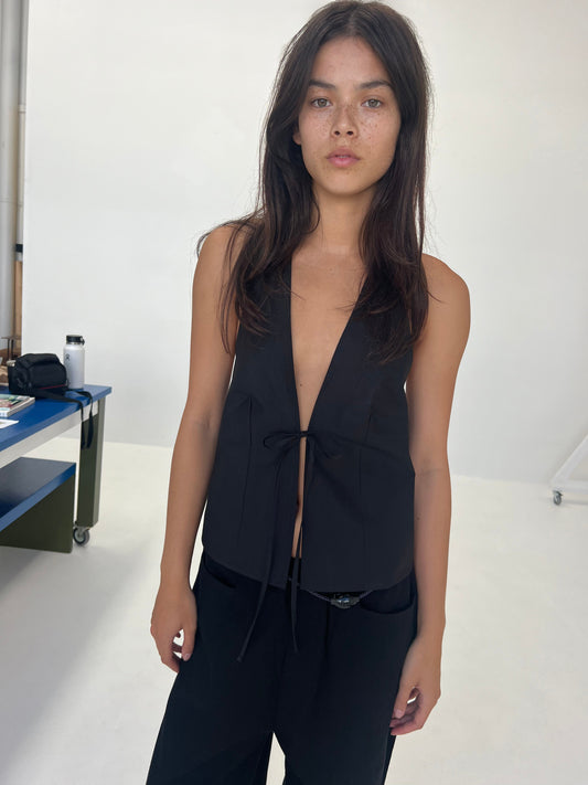 A woman with long brown hair stands against a plain white wall, wearing the Open Halter Top—sleeveless with a deep V-neck and front tie—paired with black pants. She faces the camera with a neutral expression.