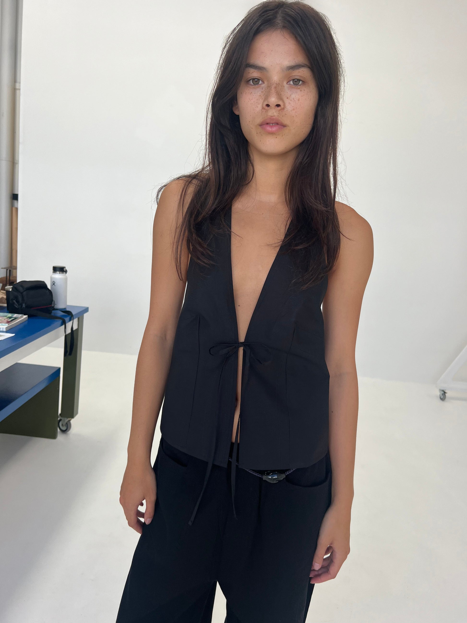 A woman with long brown hair stands against a plain white wall, wearing the Open Halter Top—sleeveless with a deep V-neck and front tie—paired with black pants. She faces the camera with a neutral expression.