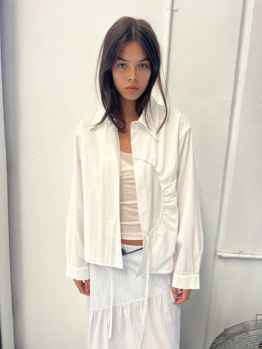 A young woman with long dark hair stands against a white wall, wearing the Arch Tie Shirt - White by Deiji Studios in organic cotton over a beige tank top and a white pleated skirt, gazing neutrally at the camera.