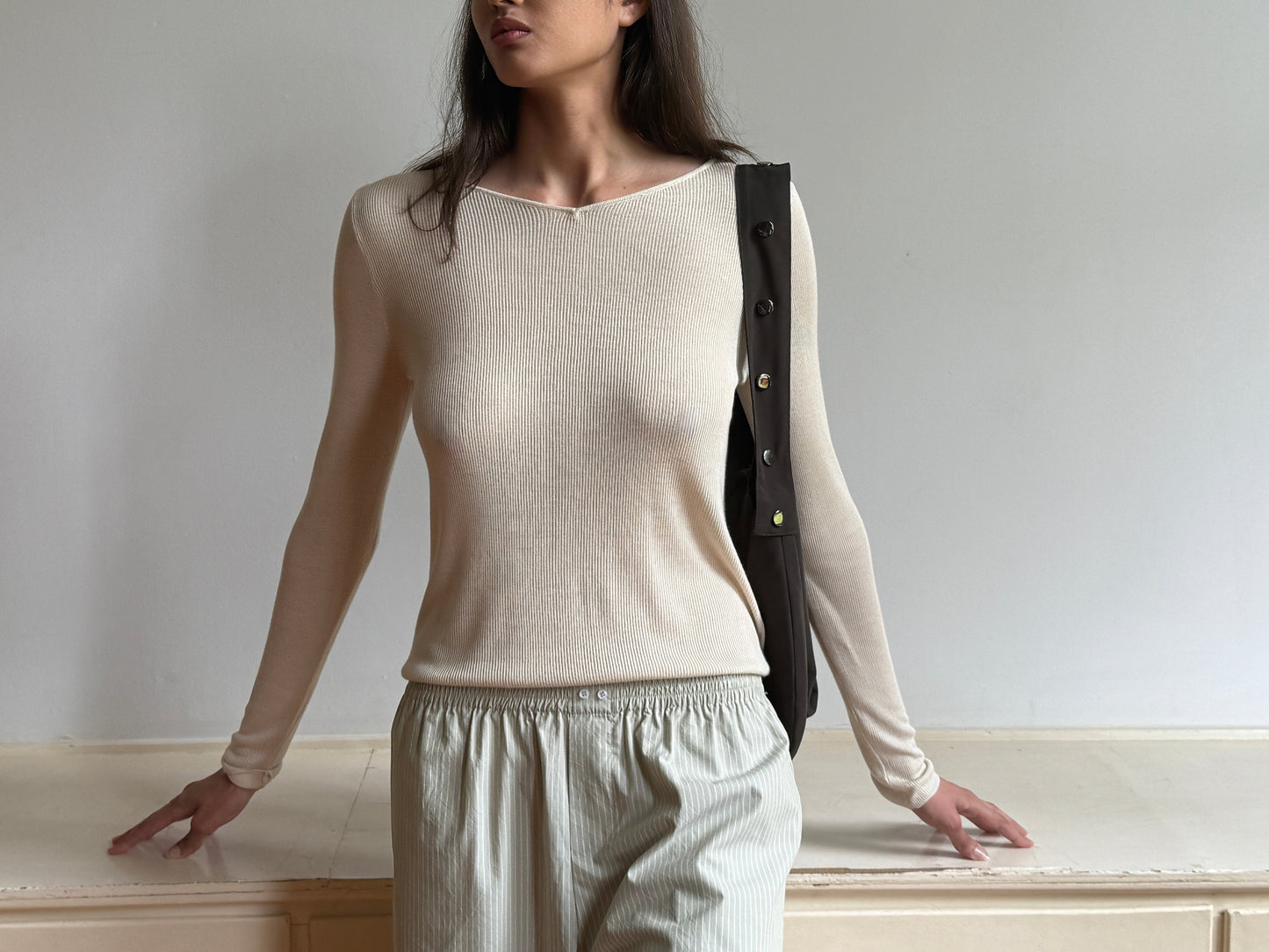 A woman stands against a white background, looking to the side, wearing the 0 Knitted Top - Fawn with light-colored pants and a dark shoulder bag on one shoulder.