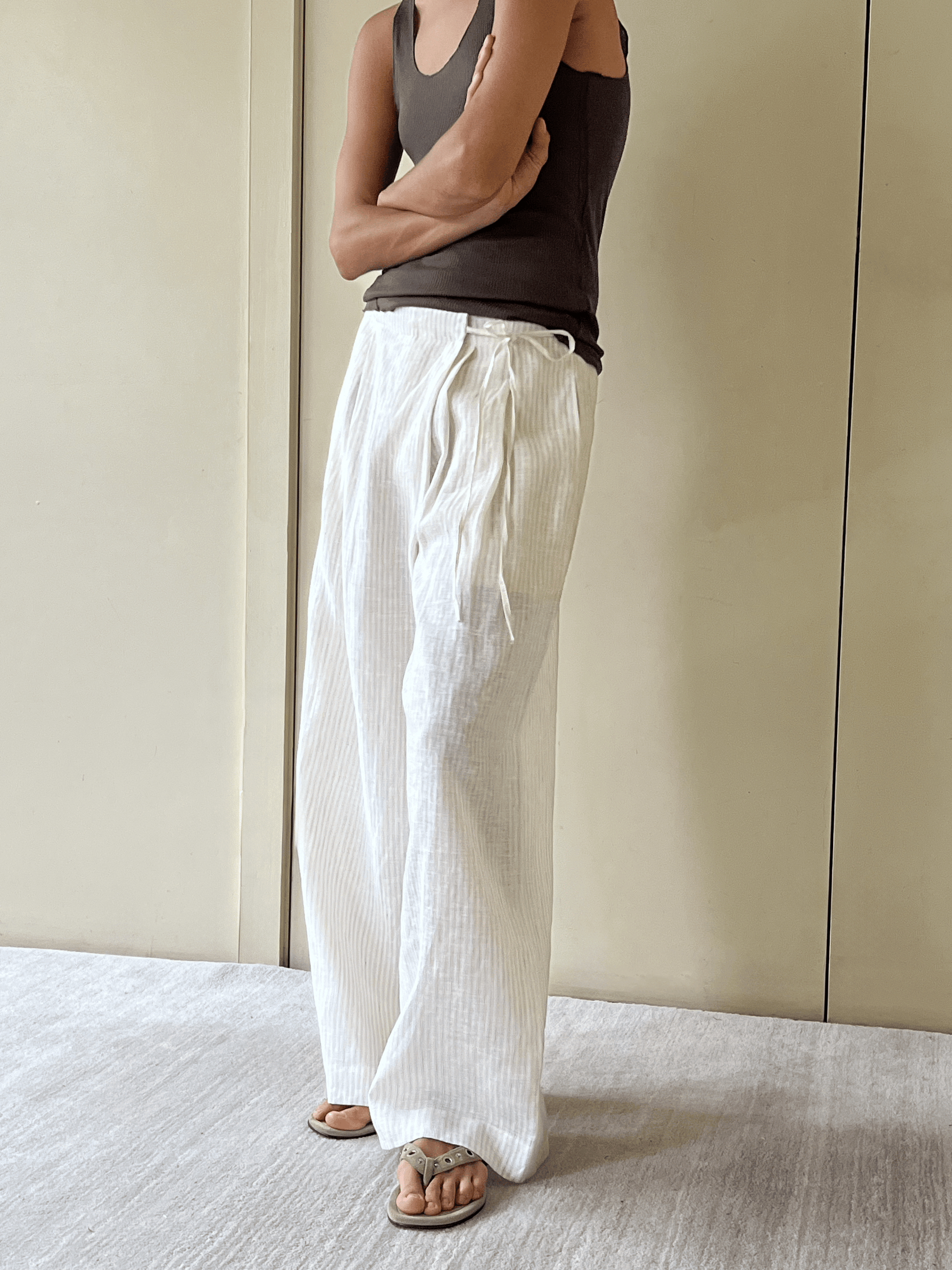 Person standing indoors with arms crossed, wearing a sleeveless dark top, the Fold Pant in white with drawstring waist, and sandals. Background shows a beige wall and a light-colored carpeted floor.