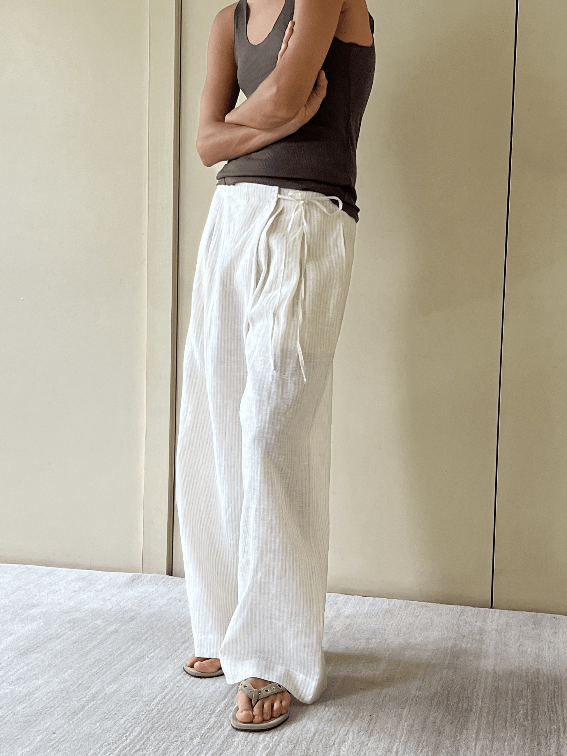 A person stands indoors with arms crossed, wearing a dark sleeveless top and the Fold Pant - Linen Story Stripe: loose, striped linen drawstring trousers. They pair this look with sandals against a neutral background of light walls and carpet.