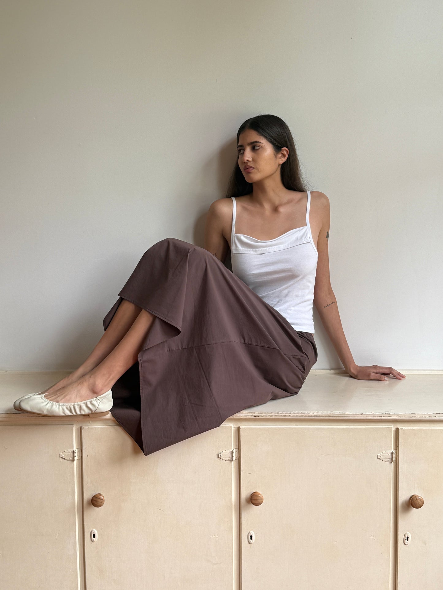 A woman with long dark hair, wearing the 0 Simple Tank in White, brown wide-leg pants, and cream flats, sits on a light cabinet and gazes to her left against a plain wall.