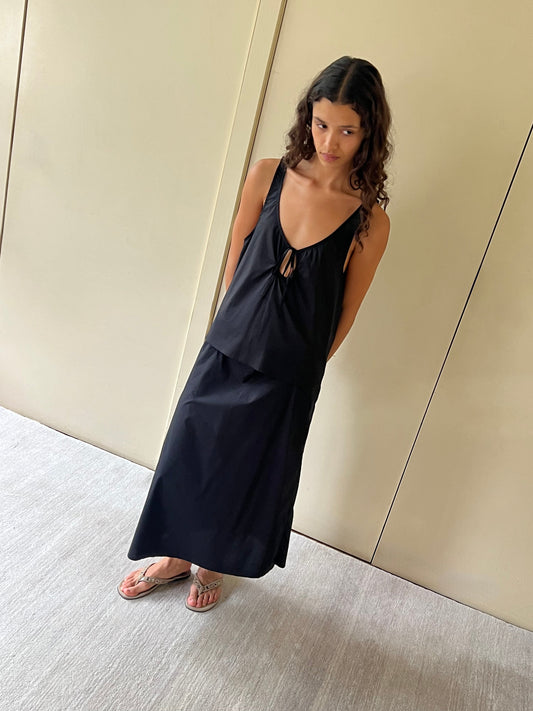 A woman with long curly hair stands indoors on a light carpet, wearing a loose black U Shape Top and matching long skirt with tan flip-flops. She looks to the side with a neutral expression.