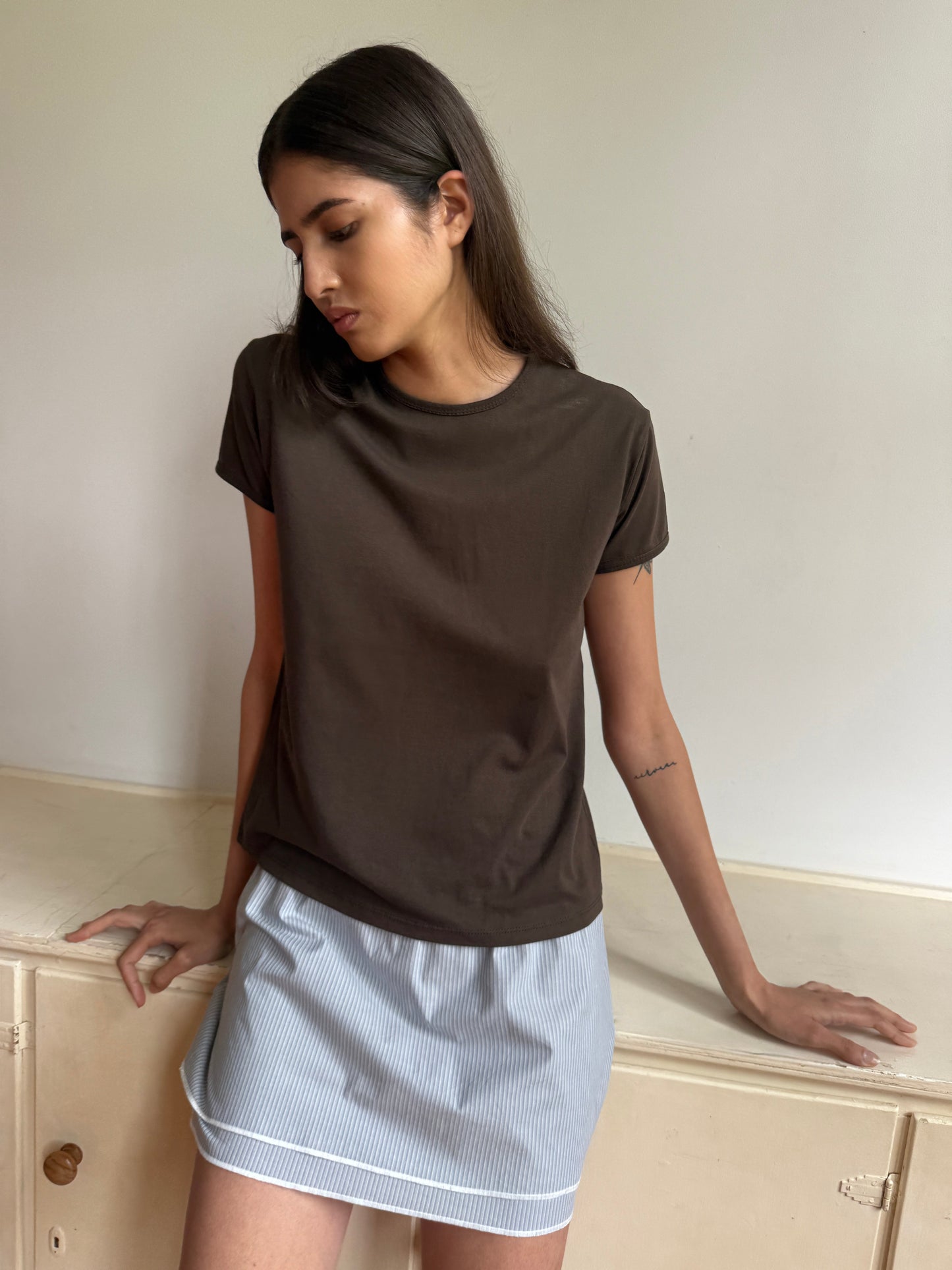 A woman with long dark hair wears the Simple Tee - Caribou by 0, paired with a light blue striped skirt, as she stands against a light-colored wall, looking down with her hands resting on a cabinet.