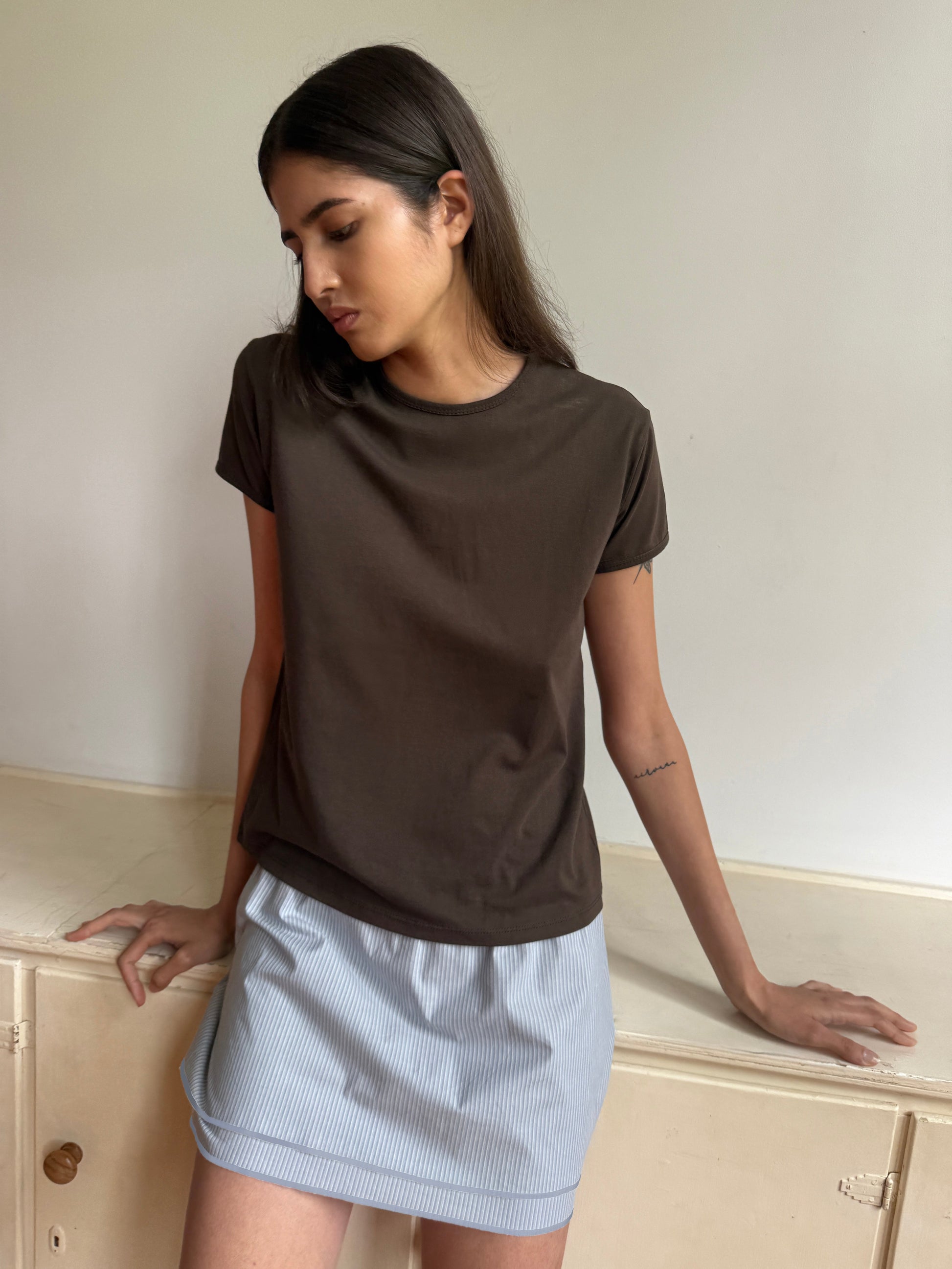 A woman with long dark hair wears a brown t-shirt and the Layered Skirt - Sky Stripe, standing indoors by a white cabinet, looking down with a neutral expression.