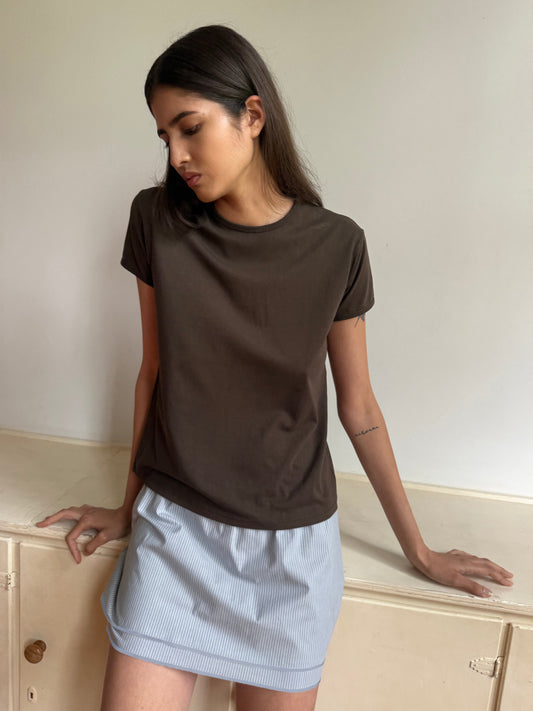 A woman with long dark hair wears a brown t-shirt and the Layered Skirt - Sky Stripe, standing indoors by a white cabinet, looking down with a neutral expression.