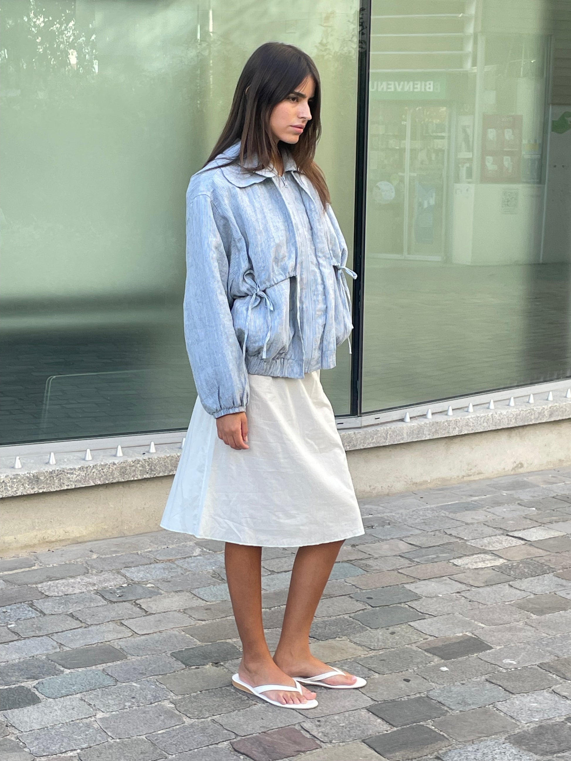 A woman stands on a stone sidewalk in an urban setting wearing the Double Layered Jacket - Blue Stripe over a knee-length white skirt and white flip-flops. She has straight, dark hair and gazes to the side.