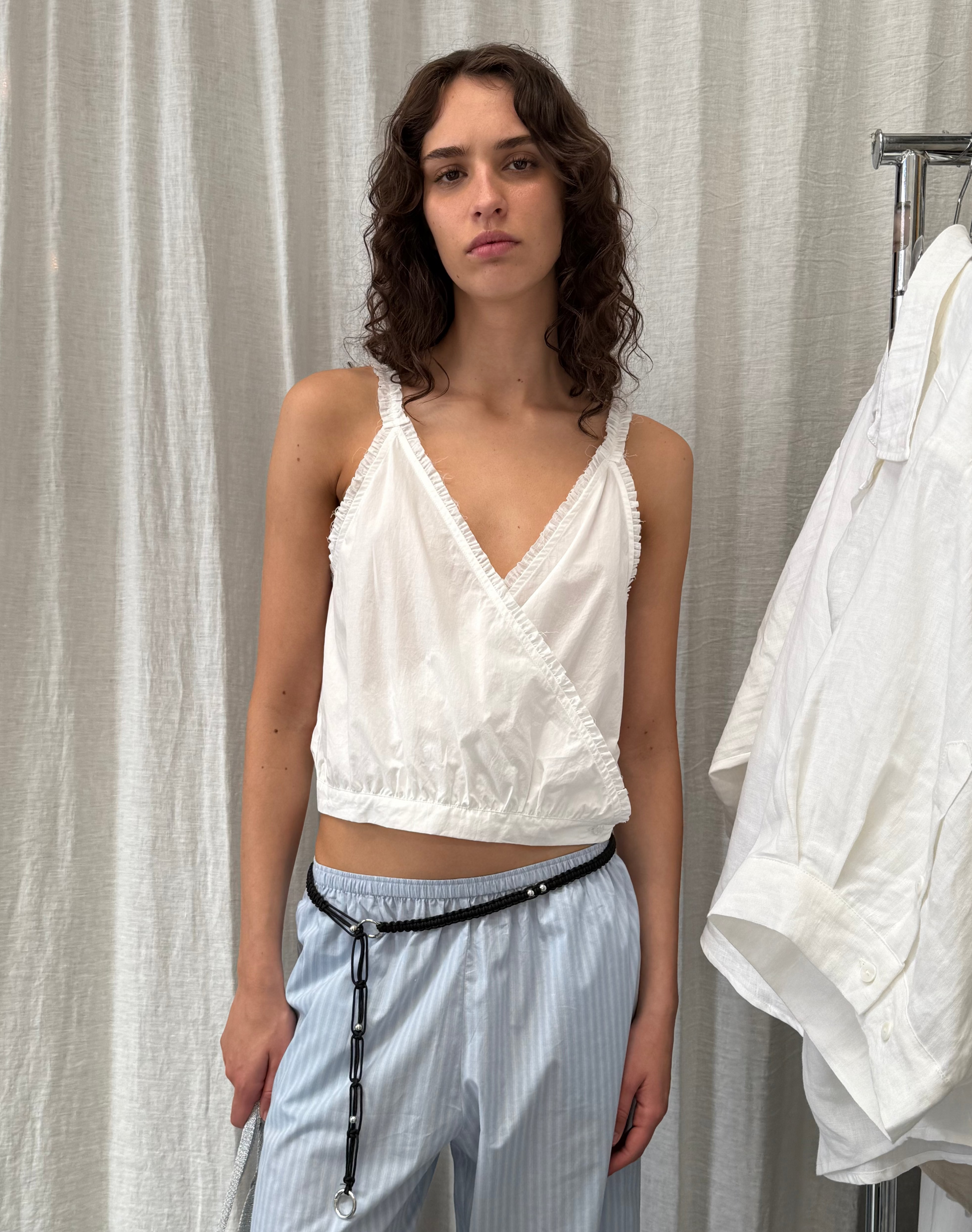 A person with wavy brown hair wears the Overlie Top - White with light blue striped pants and a thin black belt, standing against a white curtain with a white shirt hanging nearby.