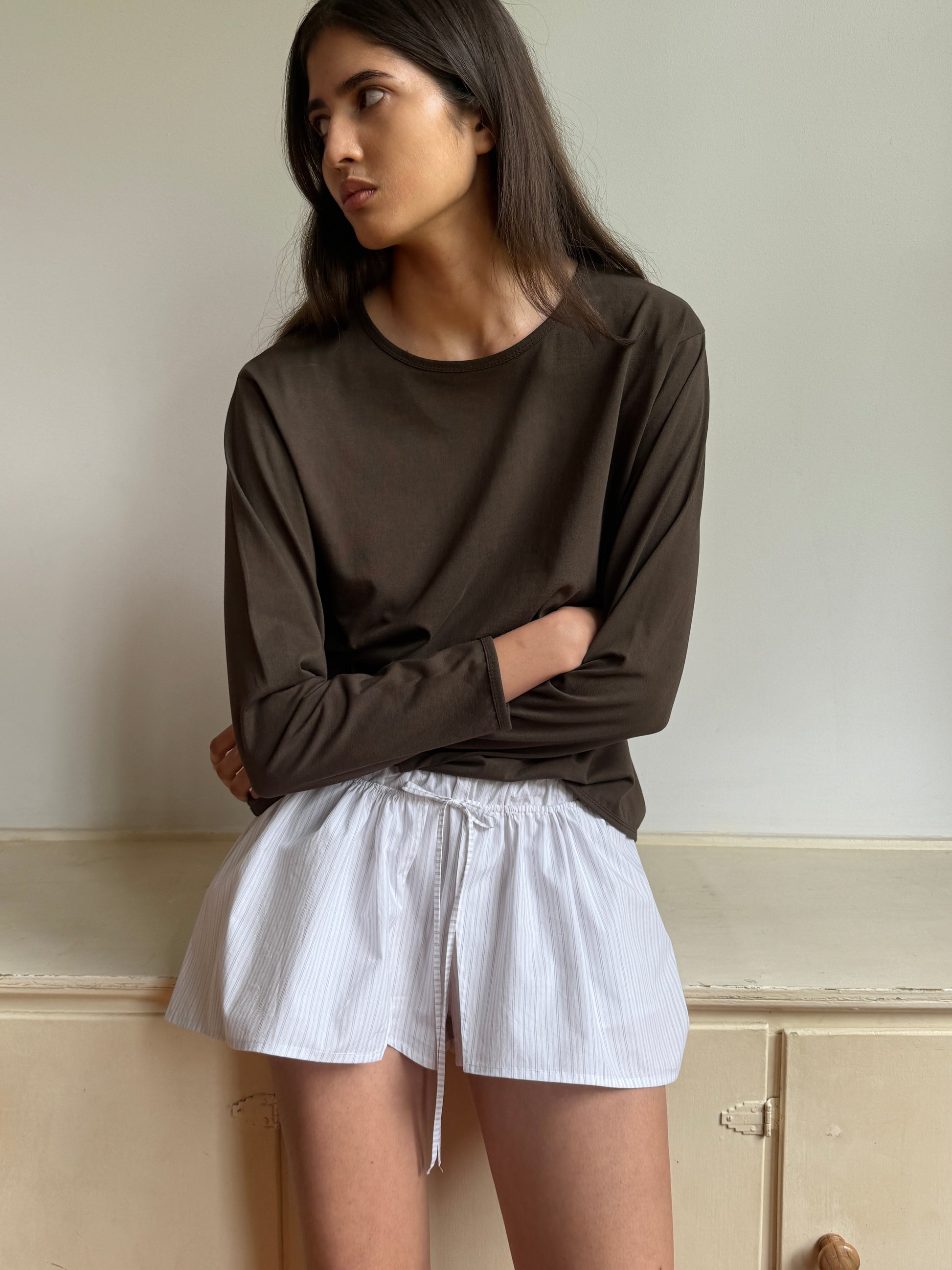 A woman with long dark hair sits on a light wooden surface, wearing the Simple Long Sleeve in dark brown and white drawstring shorts, gazing to the side against a plain, light-colored wall.